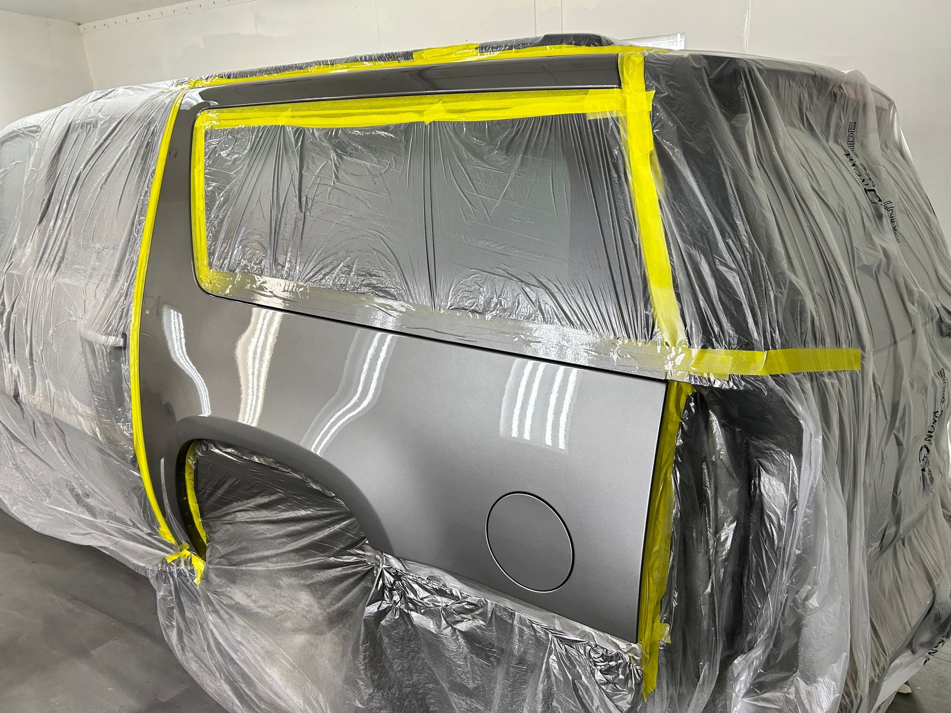 Silver SUV in a paint booth, partially covered with plastic and yellow masking tape, ready for painting.