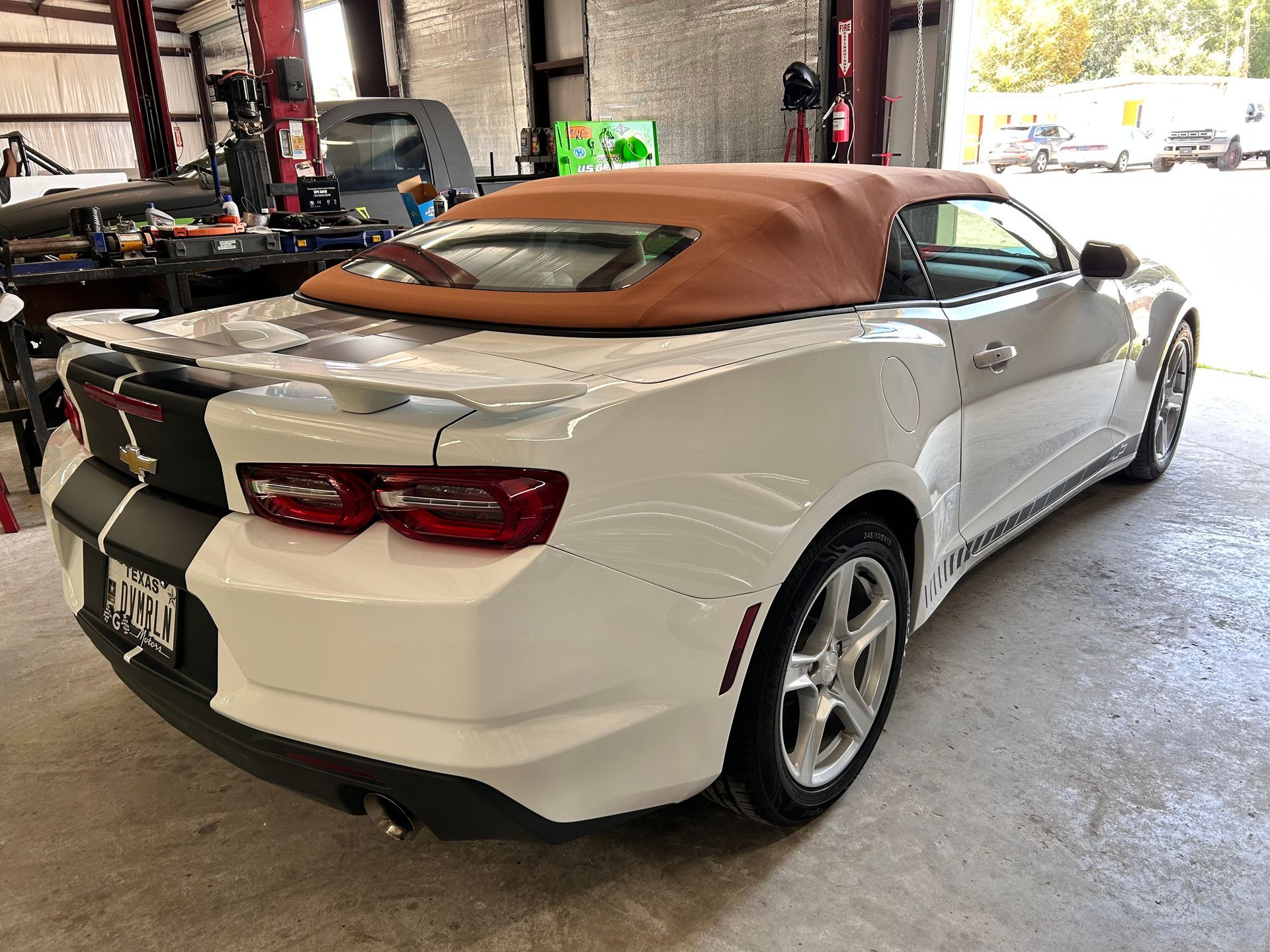 White convertible Camaro with black racing stripes and a brown top, parked indoors.