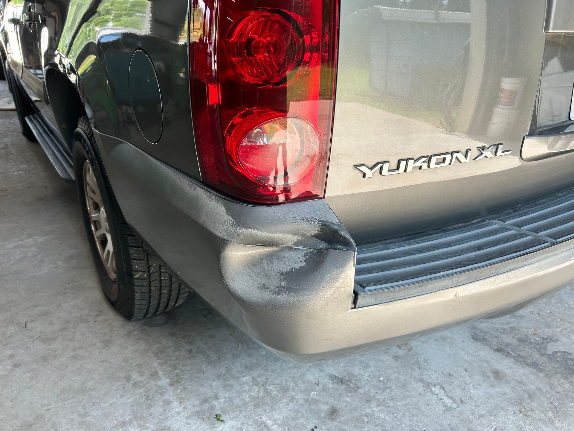 Damaged rear bumper of a tan GMC Yukon SUV. The bumper has a large dent and chipped paint.