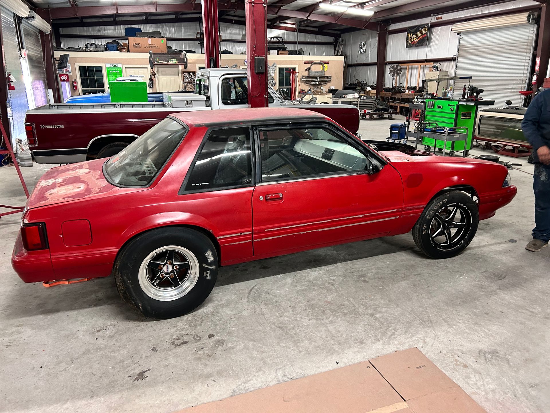 Red Ford Mustang in a garage, possibly for repairs or restoration, with black wheels.