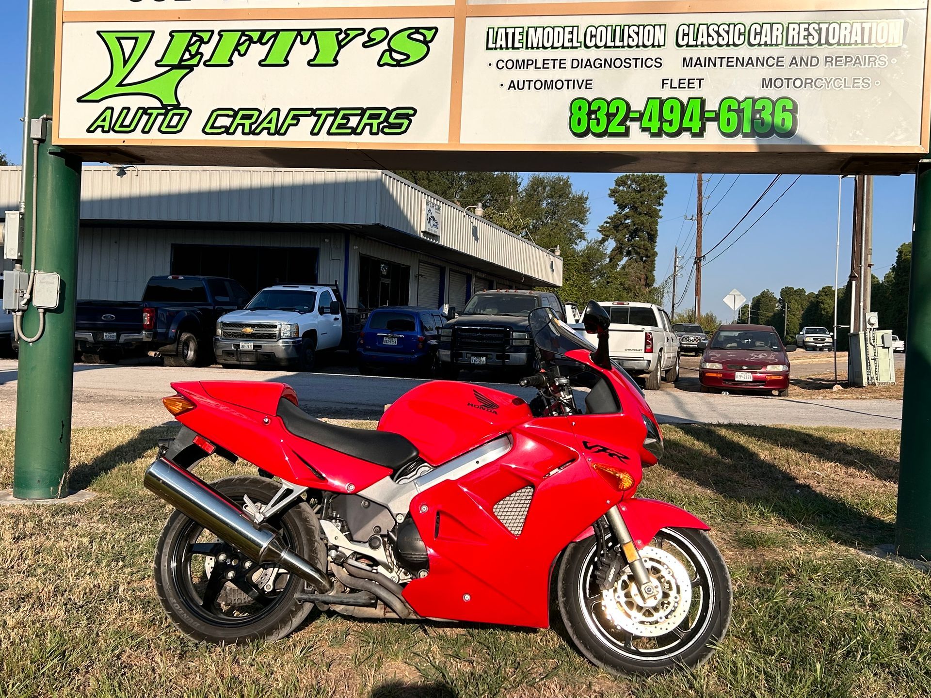 Red Honda motorcycle parked in front of an auto repair shop, sunny day.
