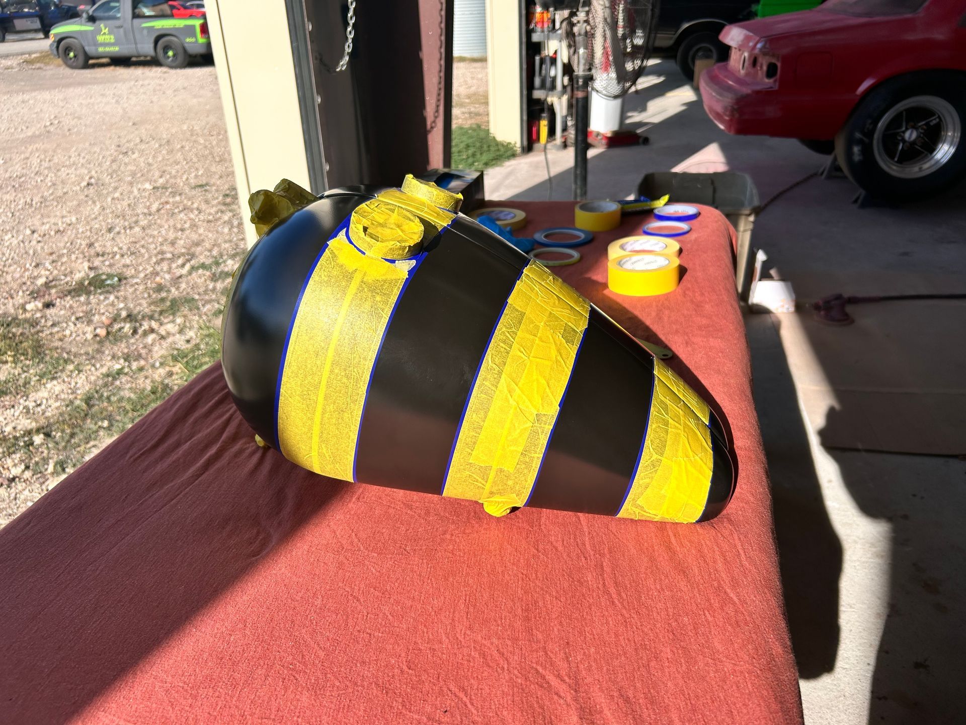 Black and yellow striped fuel tank on a red table outdoors, possibly being painted or customized.