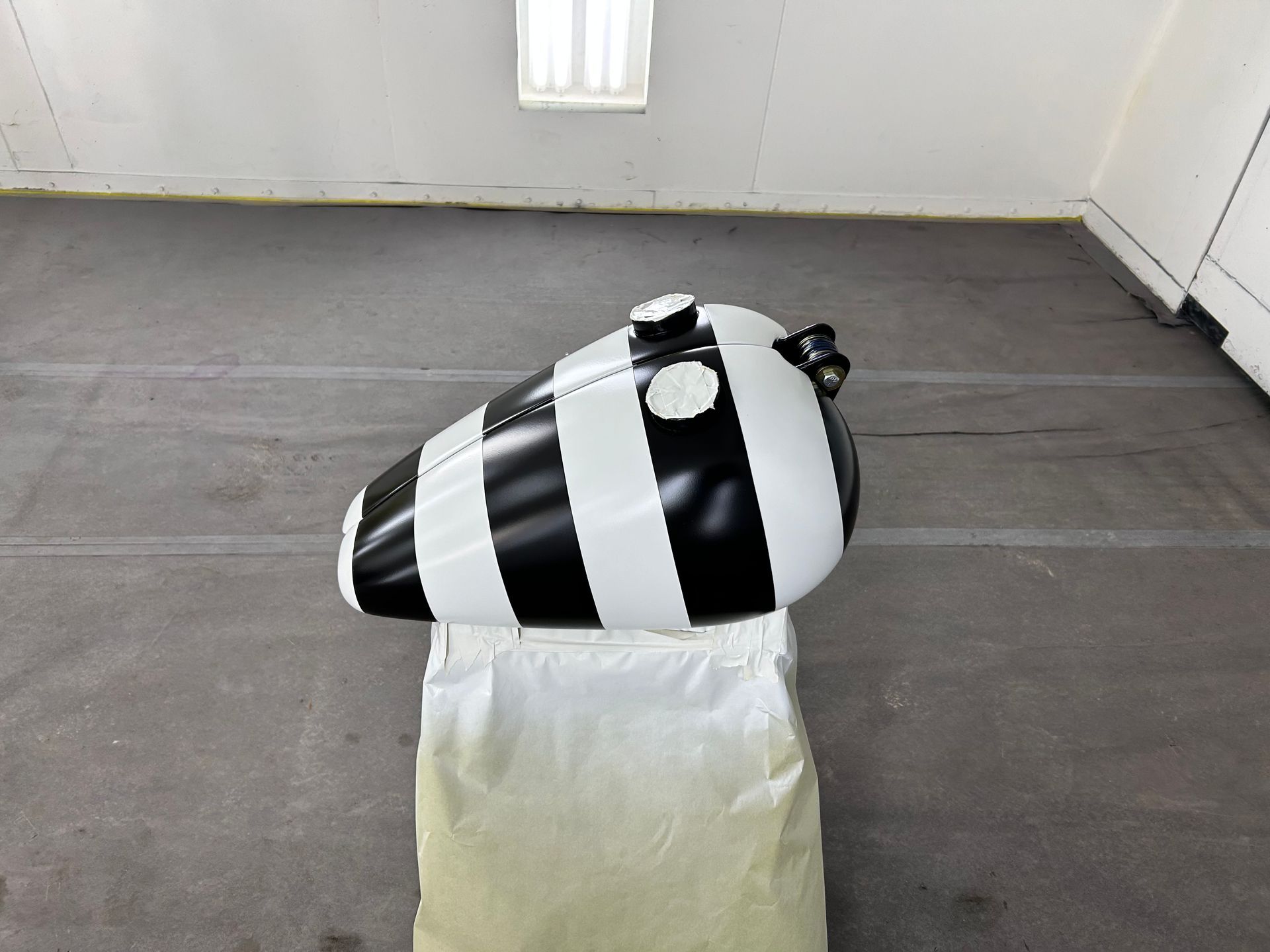 Black and white striped motorcycle gas tank on a white stand in a gray workshop.
