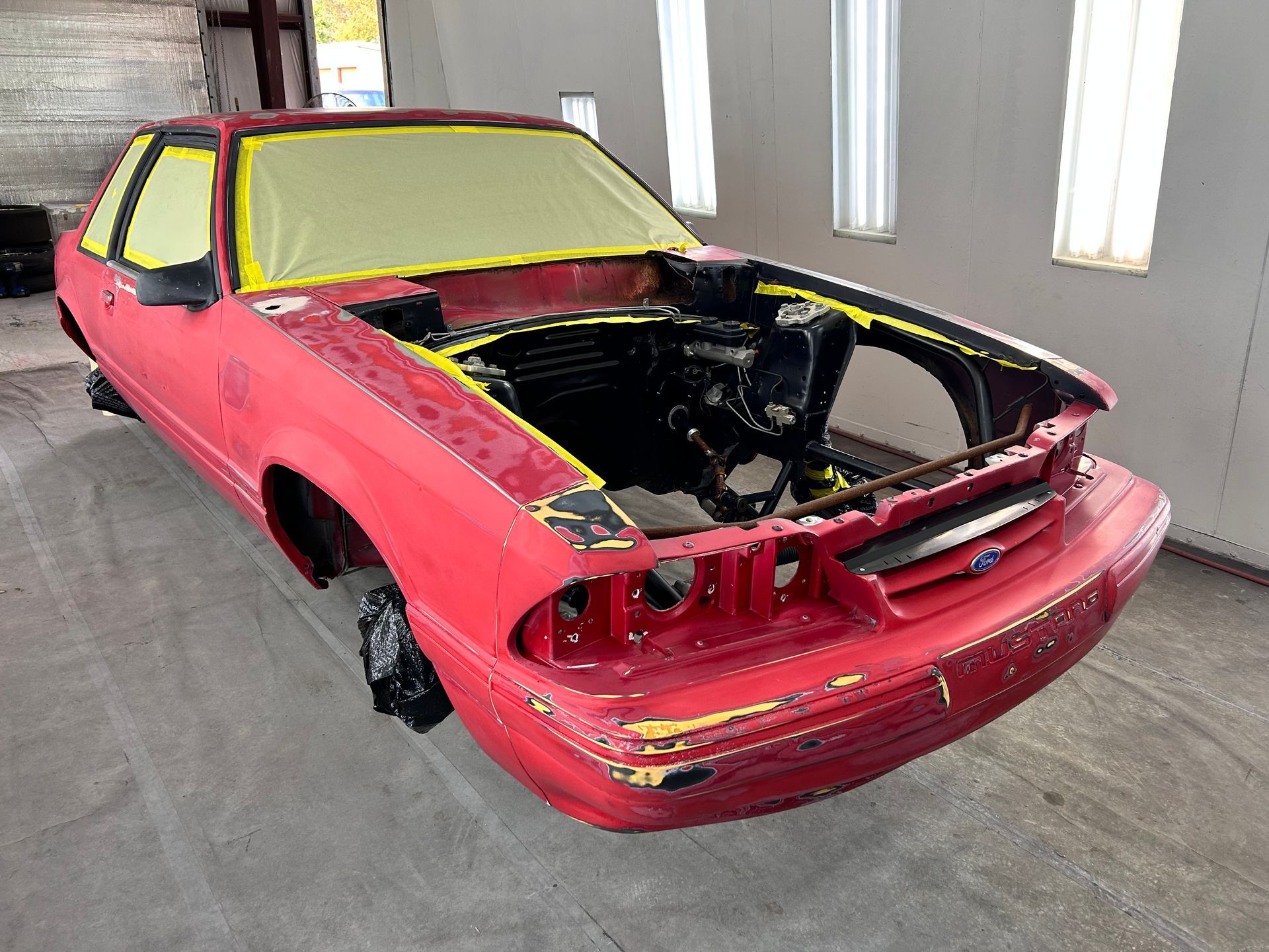 Red Ford Mustang being prepped for paint in a garage; front end stripped, yellow tape covering windows.