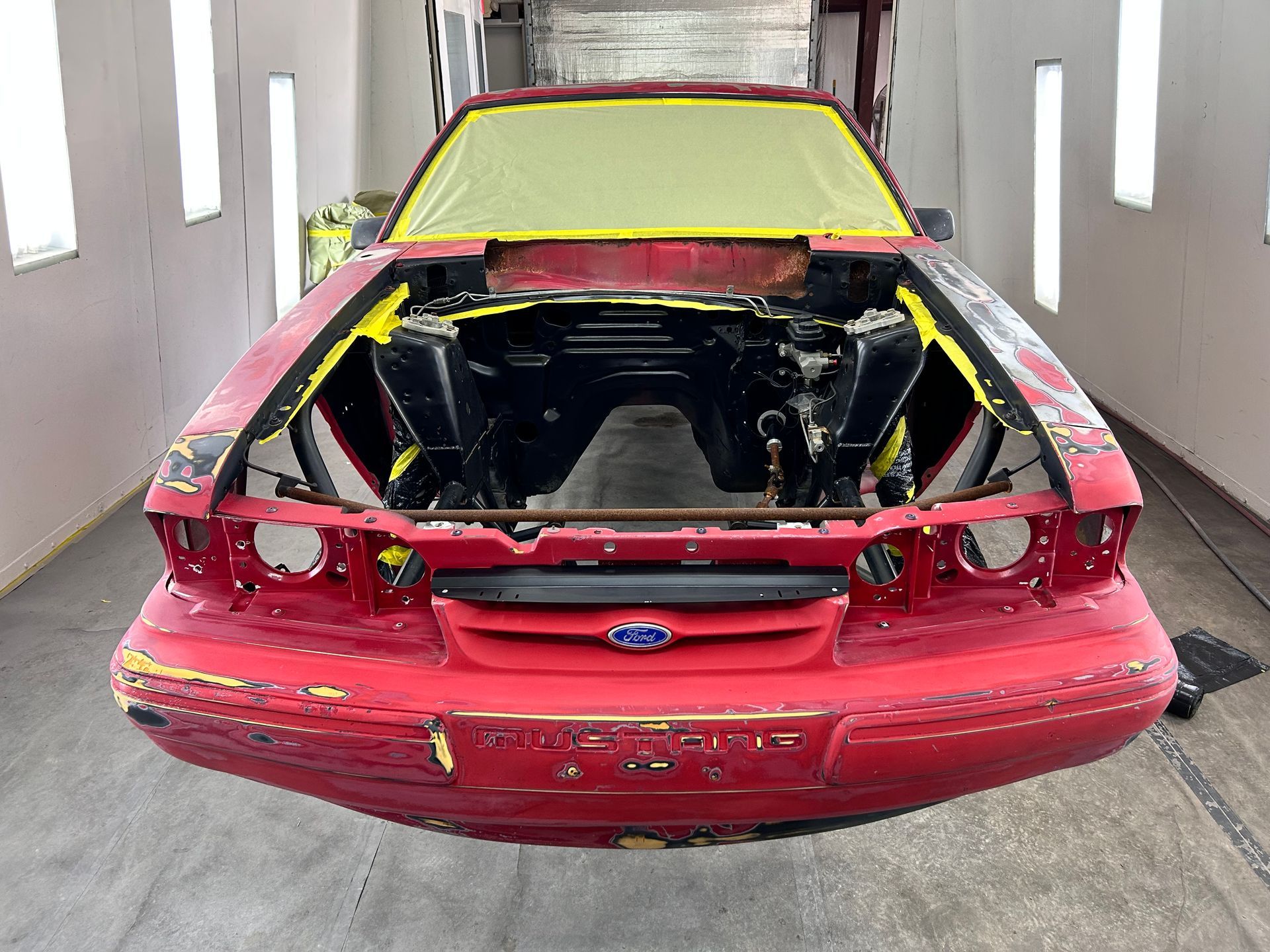 Red Ford Mustang undergoing restoration in a paint booth, masked and prepped for painting.