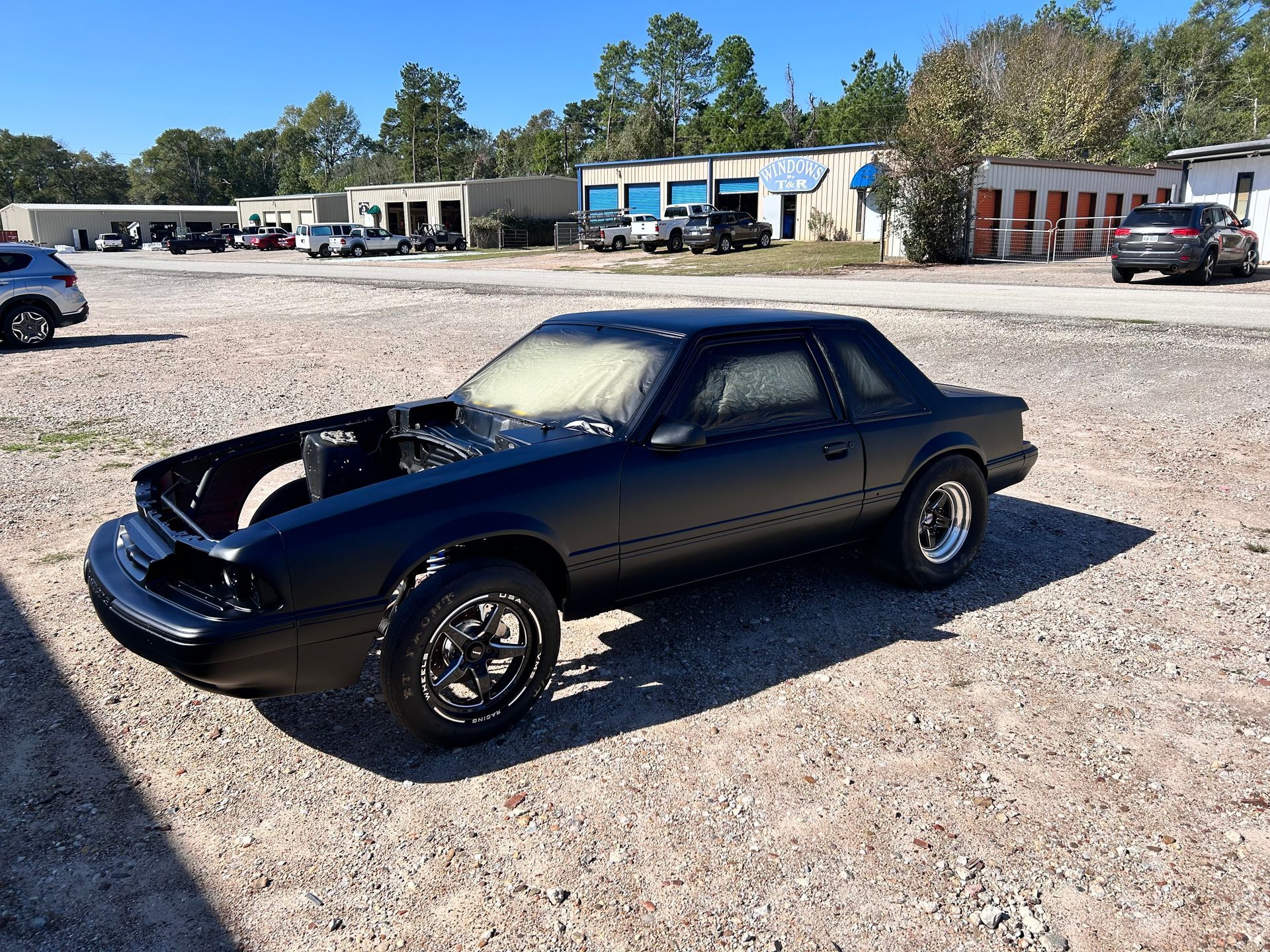 Black, unpainted Ford Mustang in a gravel lot; the front end is open, and the car is under restoration.