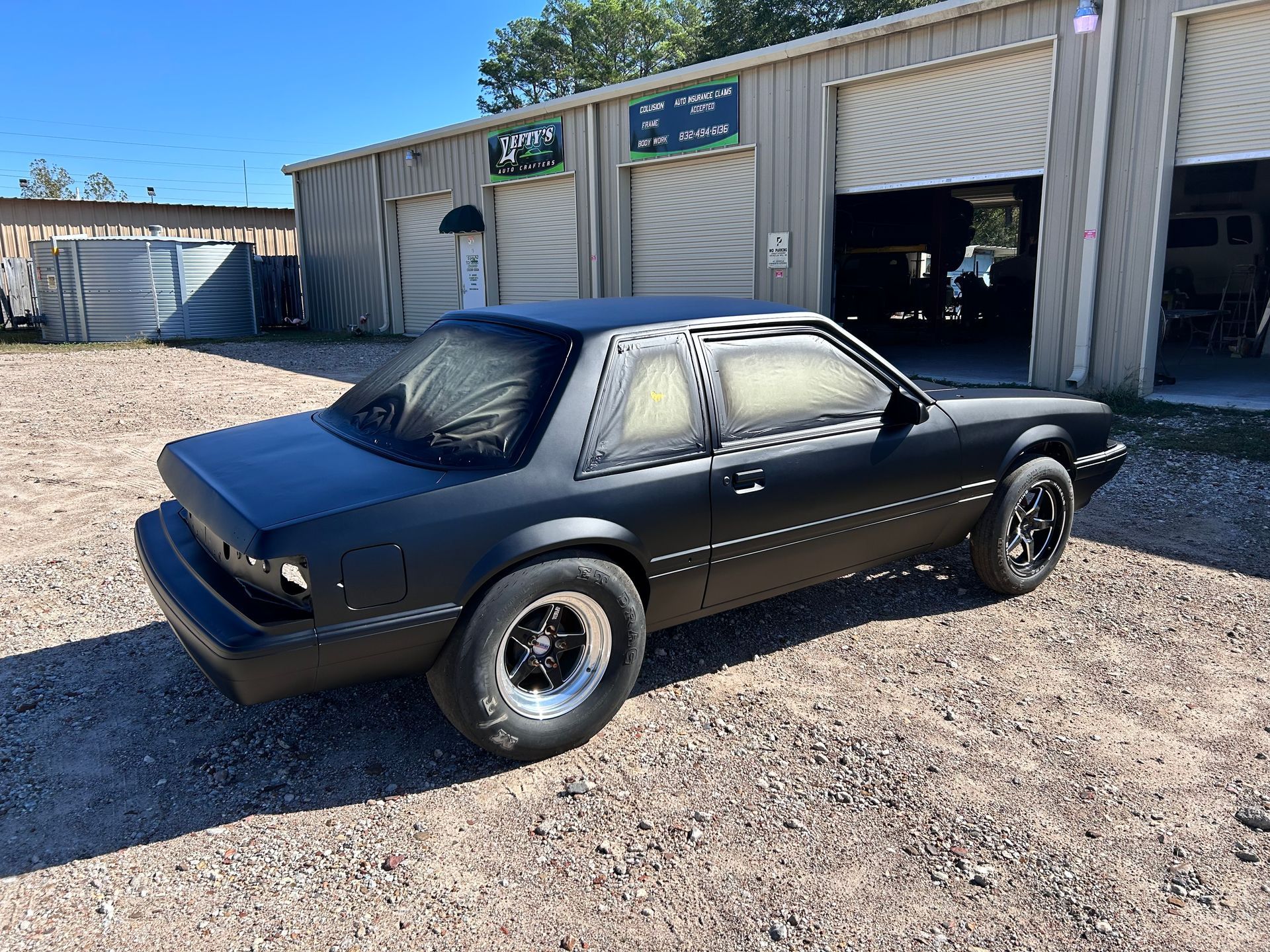Black Ford Mustang, primed for paint, parked outside a garage on a sunny day.