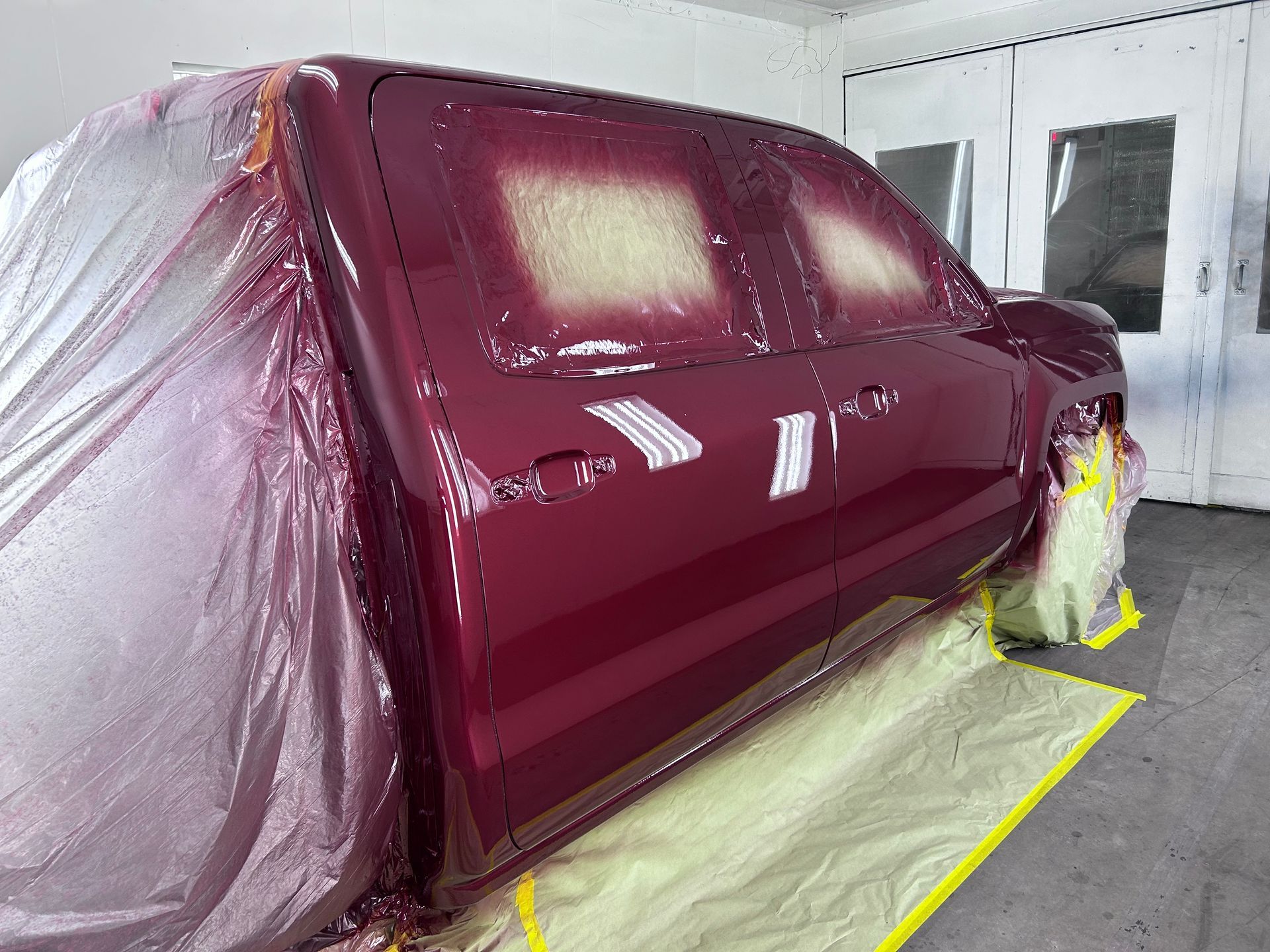 Red pickup truck in a paint booth, partially masked and freshly painted maroon.