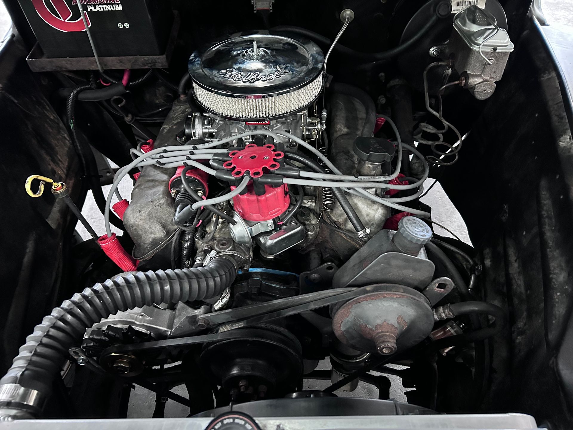 Engine compartment of a vintage car, showing the engine with a chrome air filter and red distributor cap.