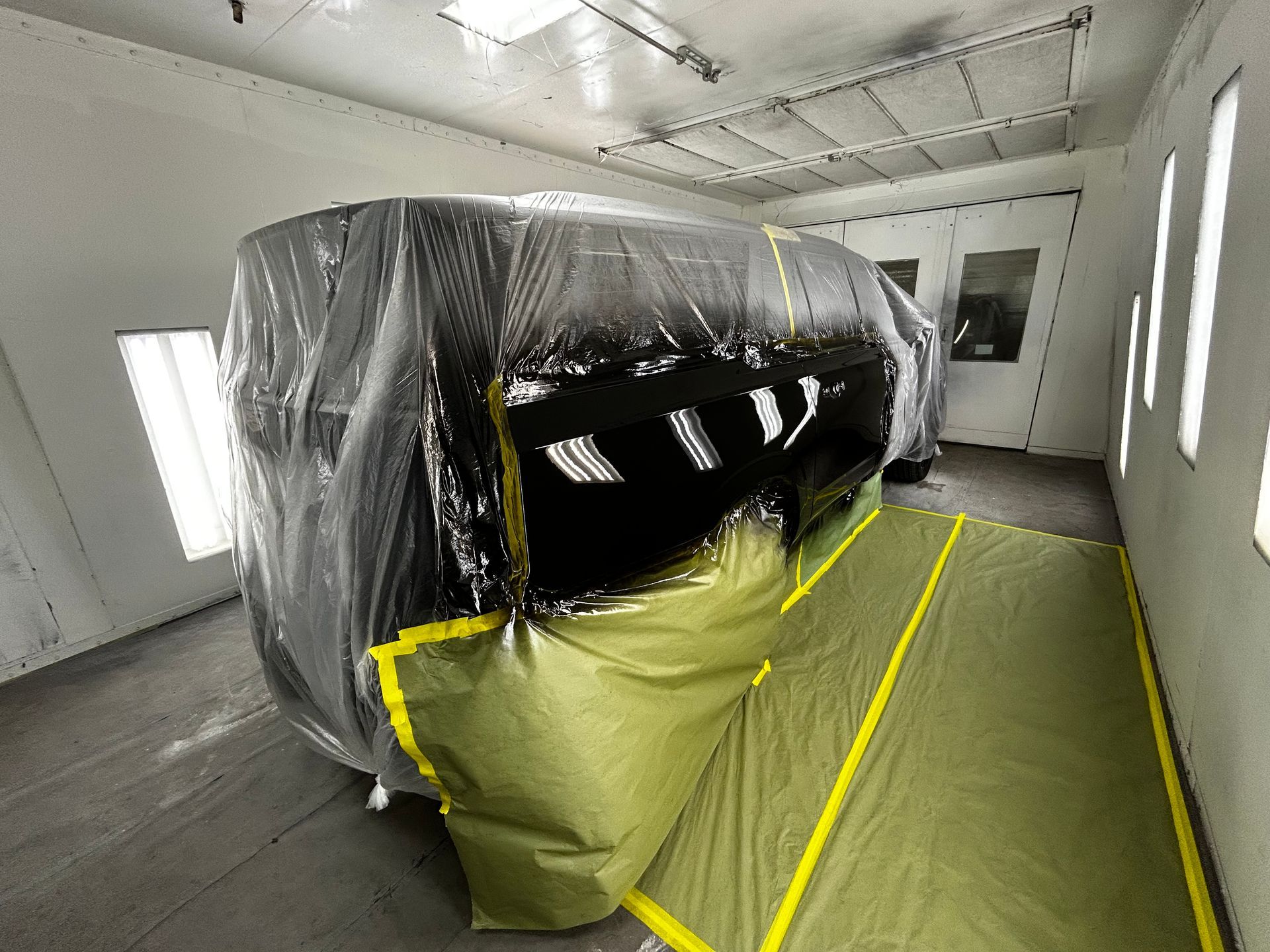 Car covered in plastic in a paint booth, masked with yellow tape, ready for paint.