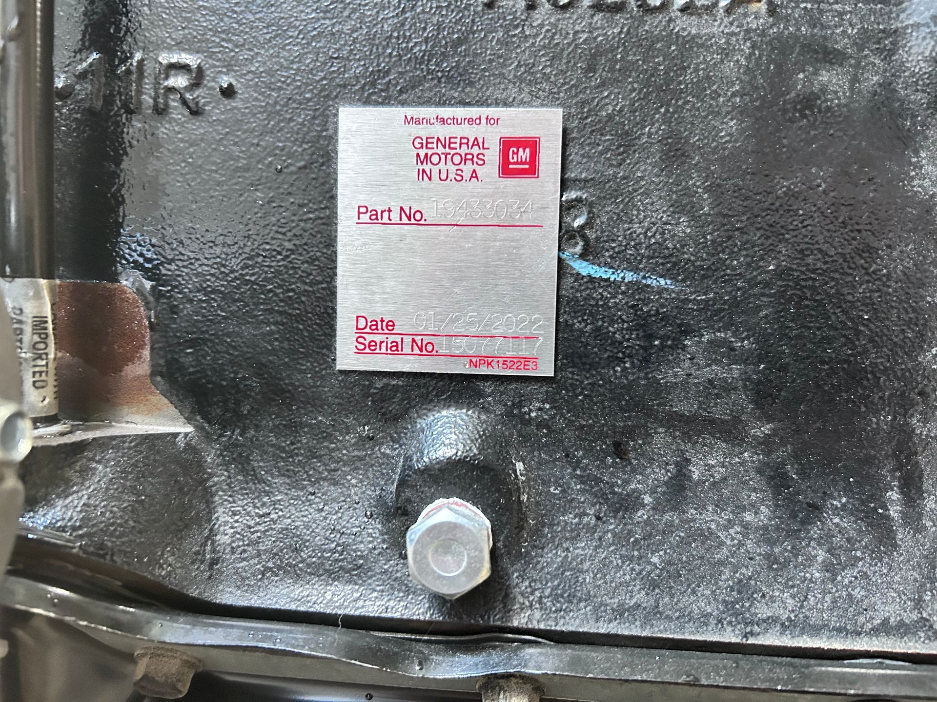Silver metal tag on a black engine block with GM logo, part and serial number fields, and 