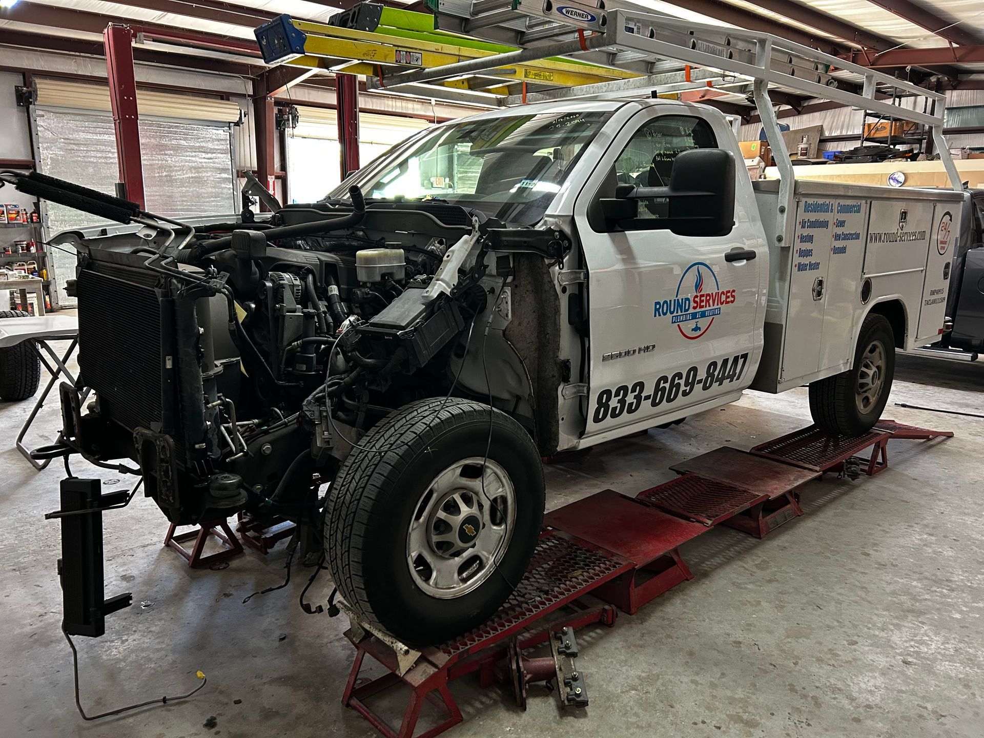 White work truck with front-end damage inside a garage, raised on a lift.
