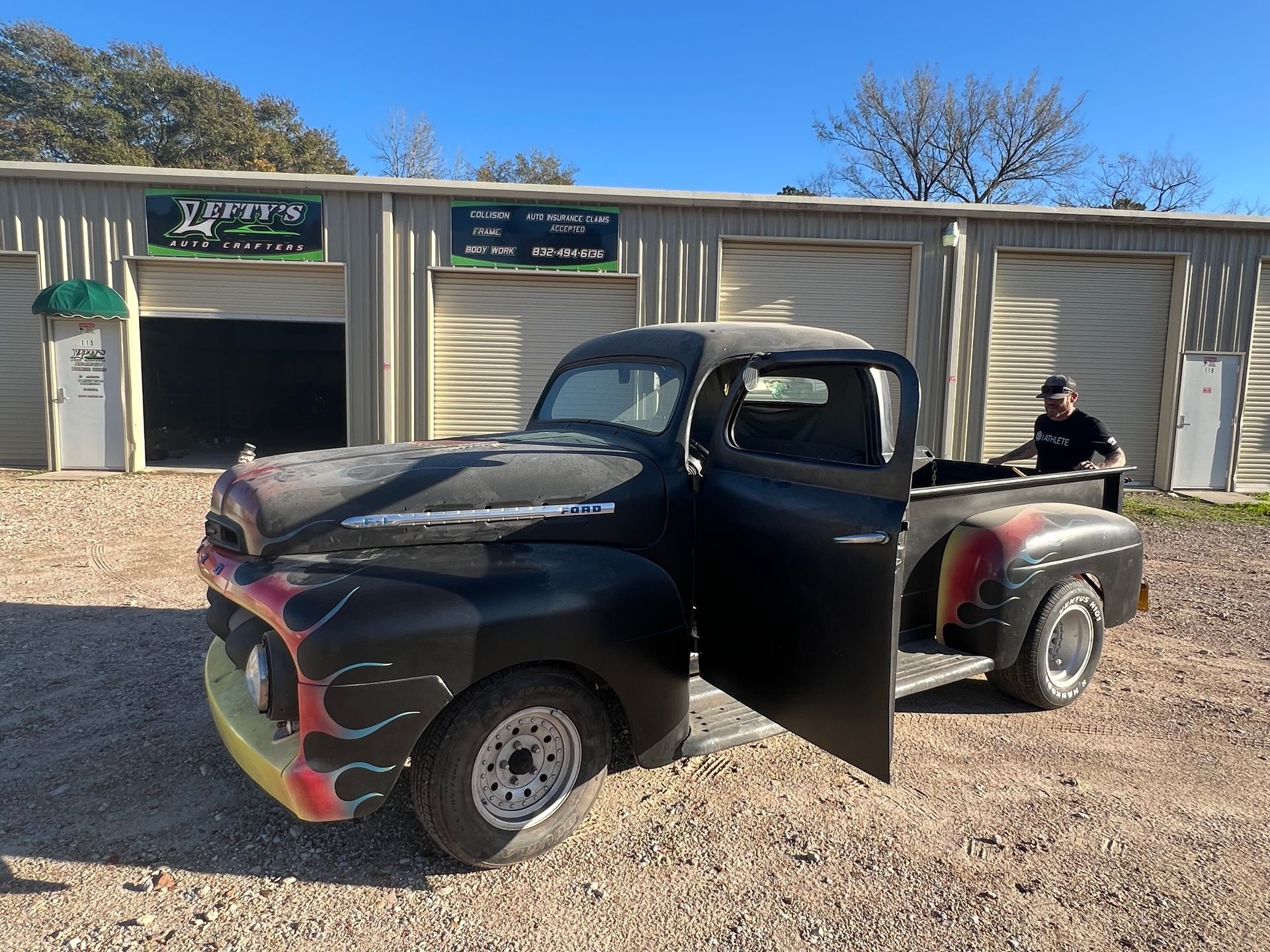 Black vintage pickup truck with flames parked in front of garage doors, door open, person visible.