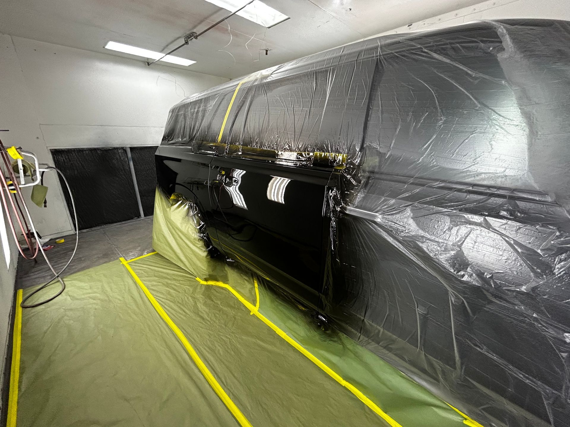 A black SUV is covered in plastic in a paint booth. Yellow tape and green mats protect the floor.