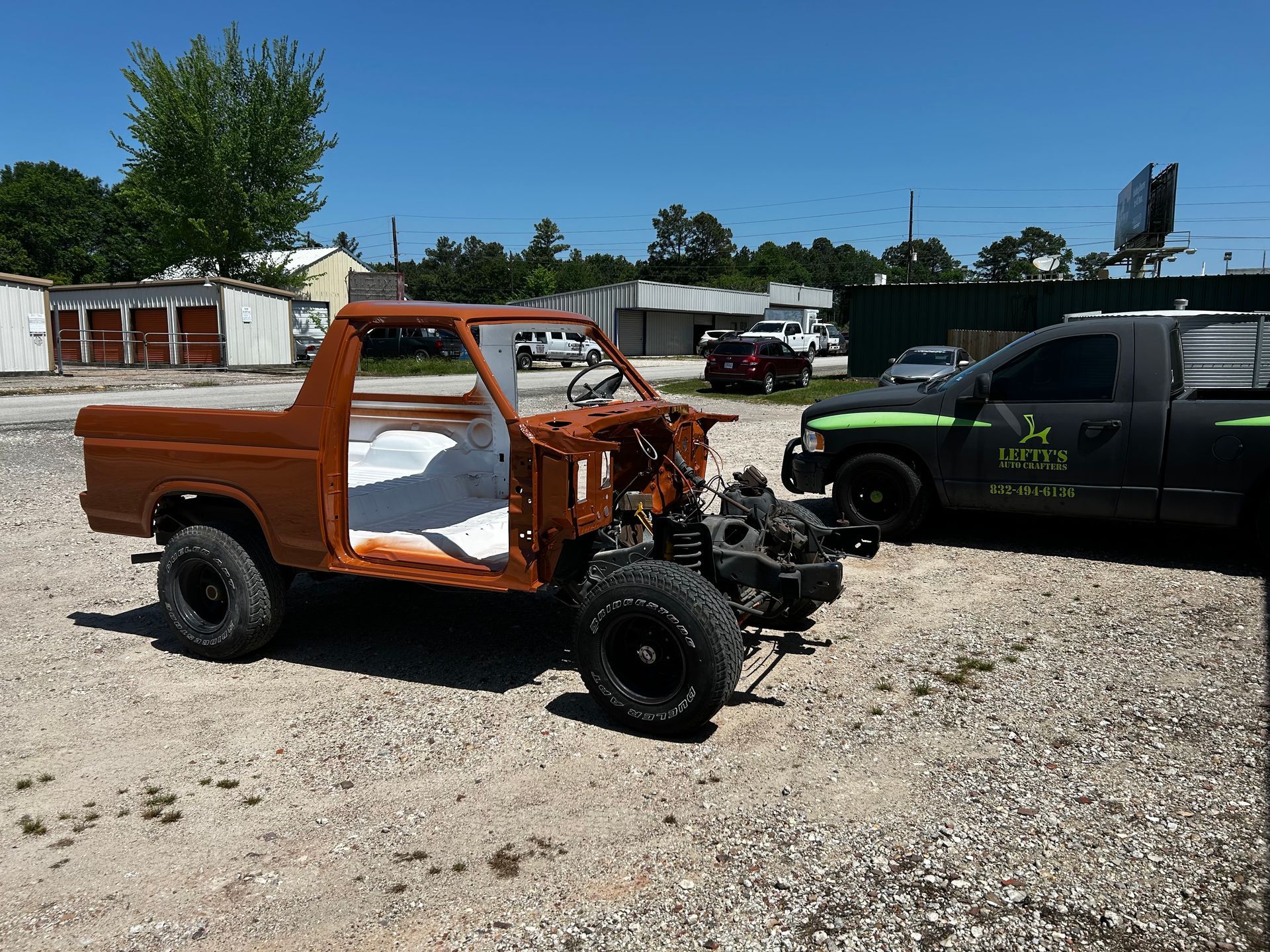 A rusty brown pickup truck body on black wheels sits on gravel in a sunny lot.