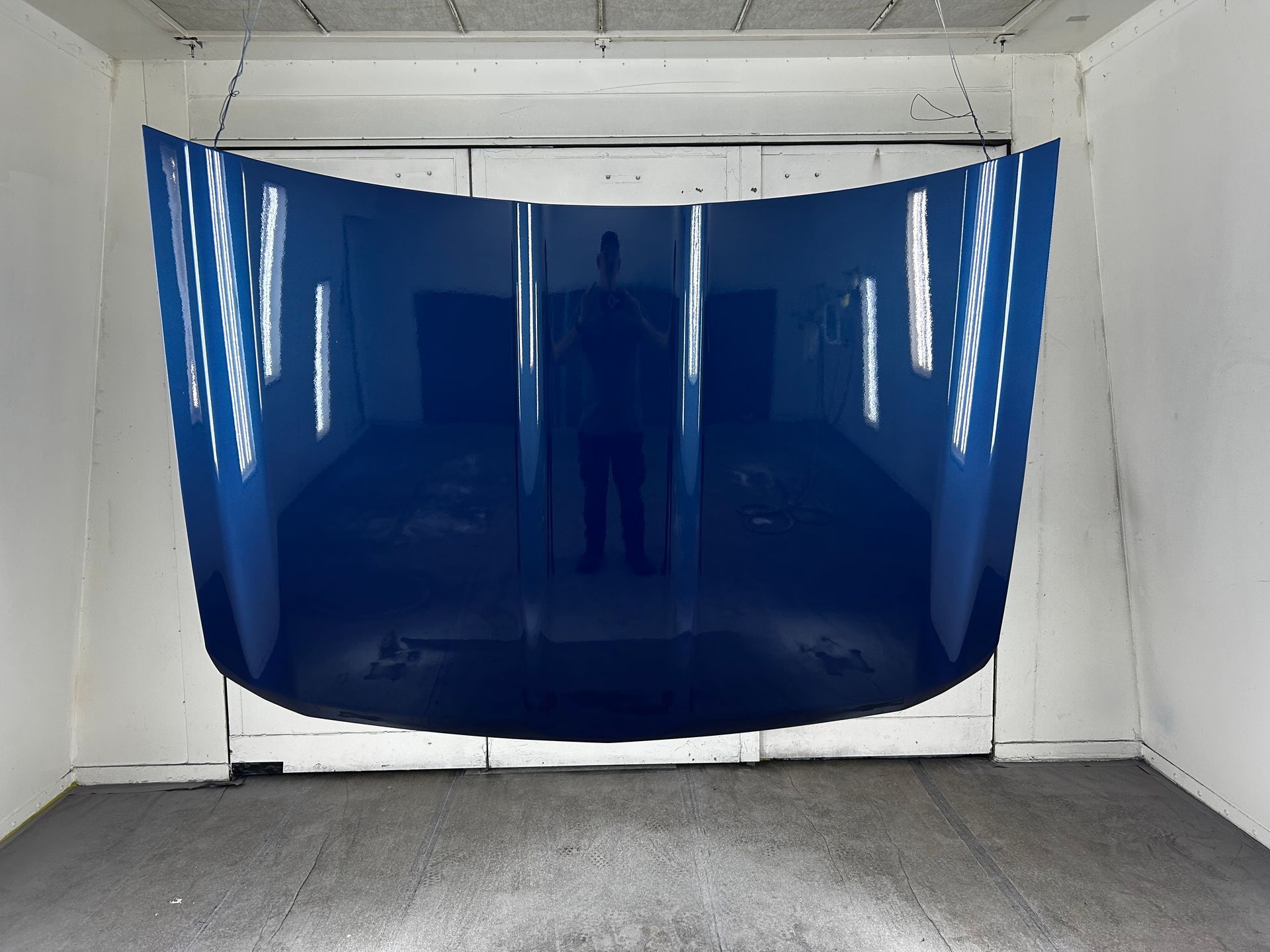 Blue car hood in a paint booth, suspended from the ceiling, with reflections of the room and person.