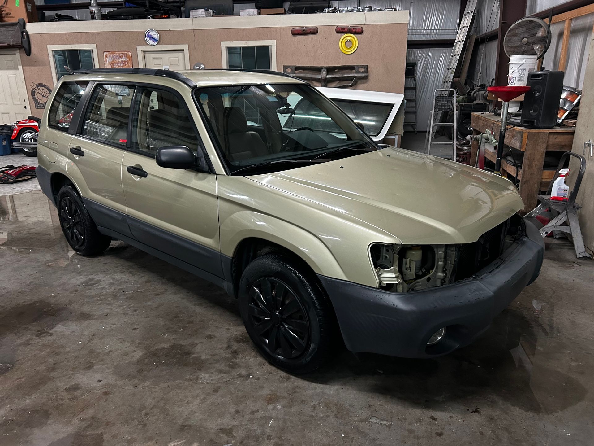 Gold Subaru Forester in a garage, front view. Black wheels, missing headlights, gray trim.