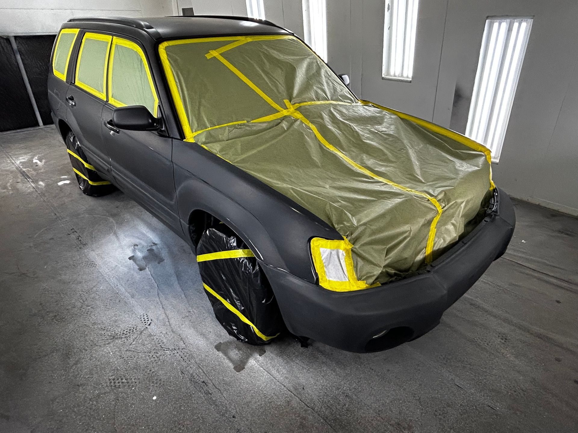 Black SUV masked with yellow tape and plastic in a paint booth.