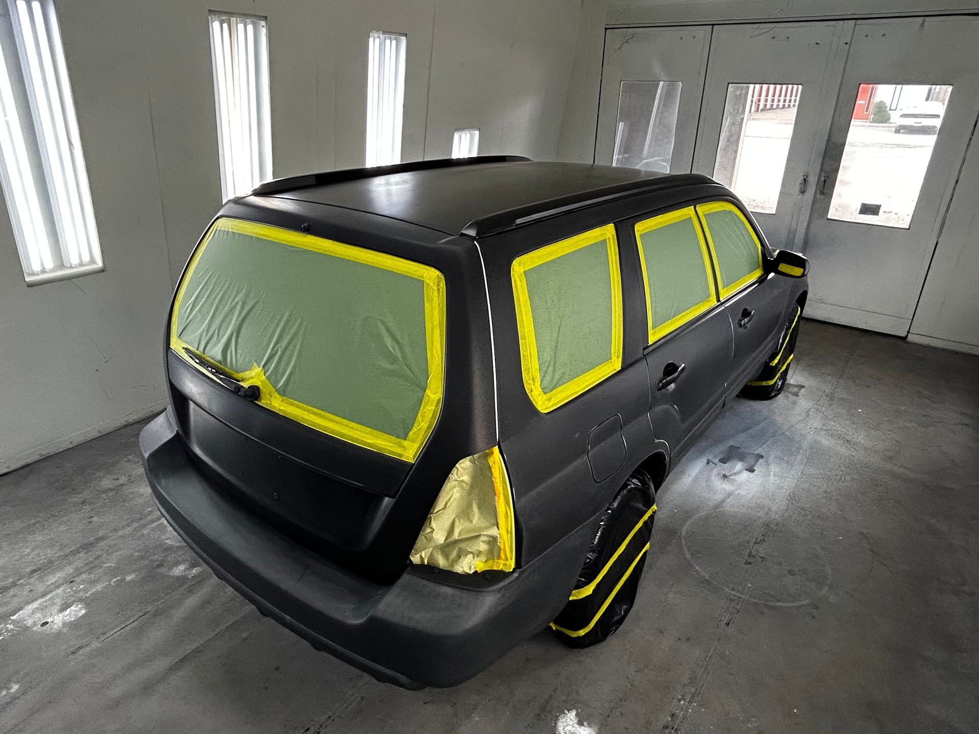 Black car taped for paint in a spray booth, windows and wheels masked.