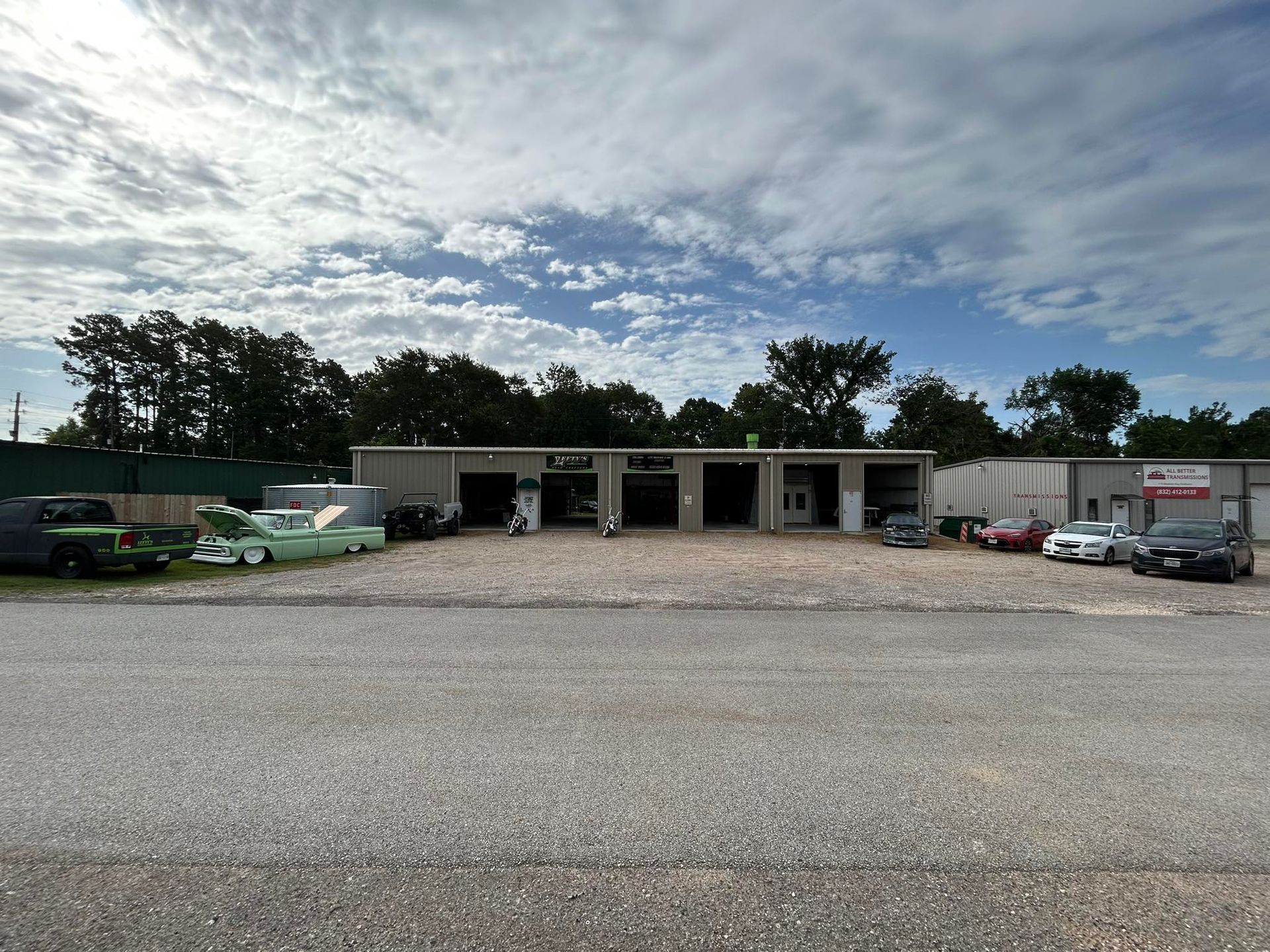 Auto repair shop with open garage bays, cars parked outside, gravel lot, cloudy sky.