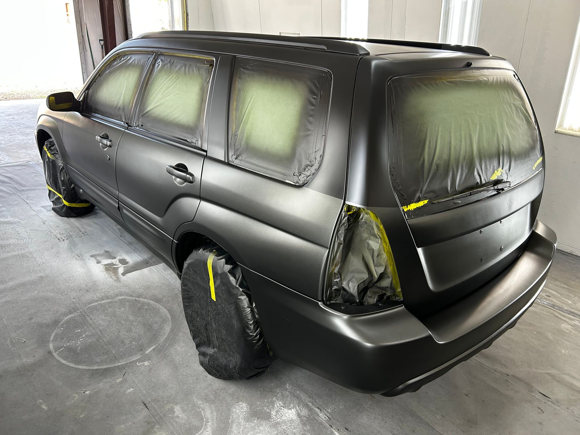 Subaru Forester in matte black paint, masked for spray, in a workshop setting.