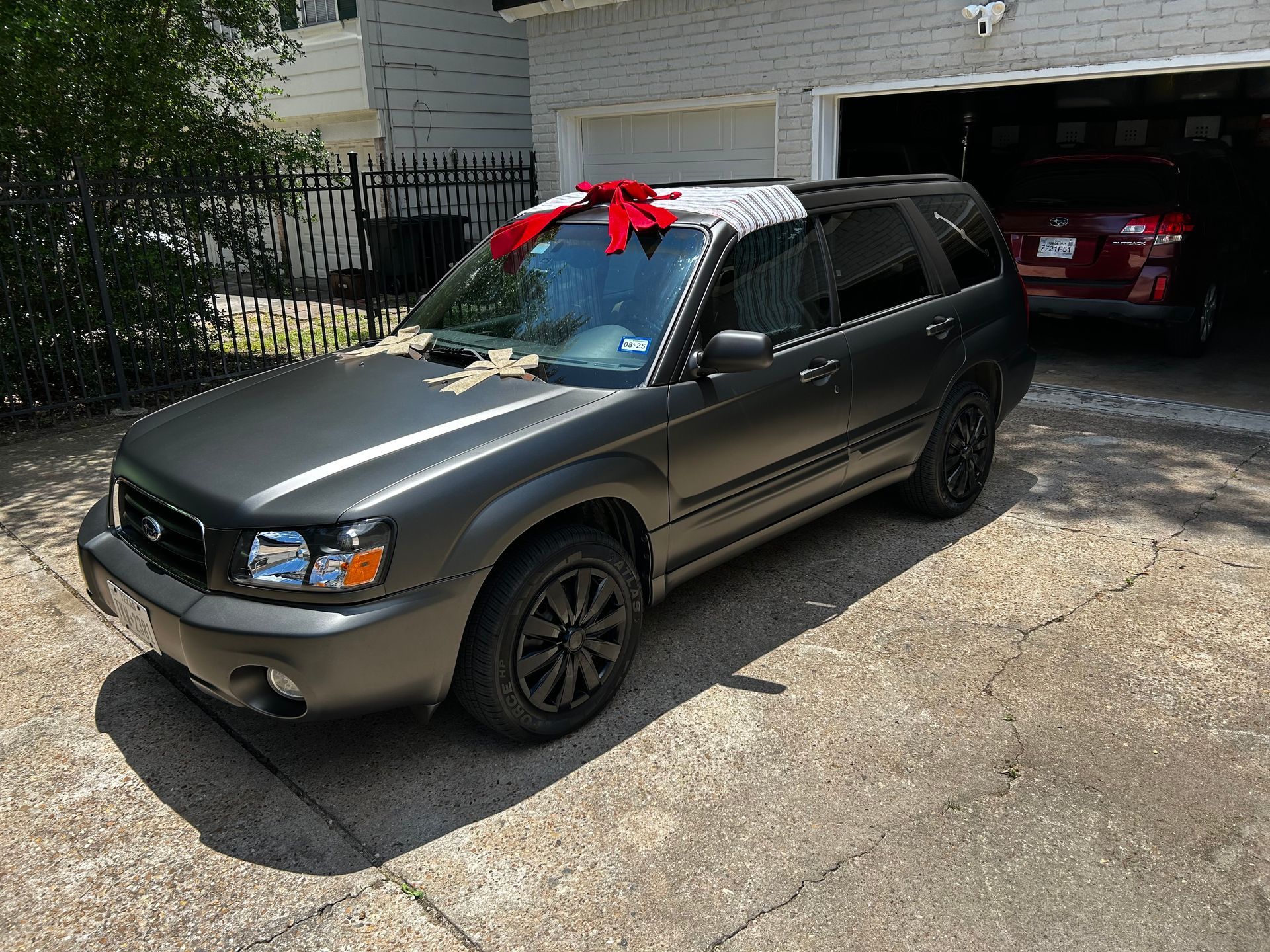 Gray Subaru Forester parked in driveway with a red bow on the roof, near a black fence and garage.
