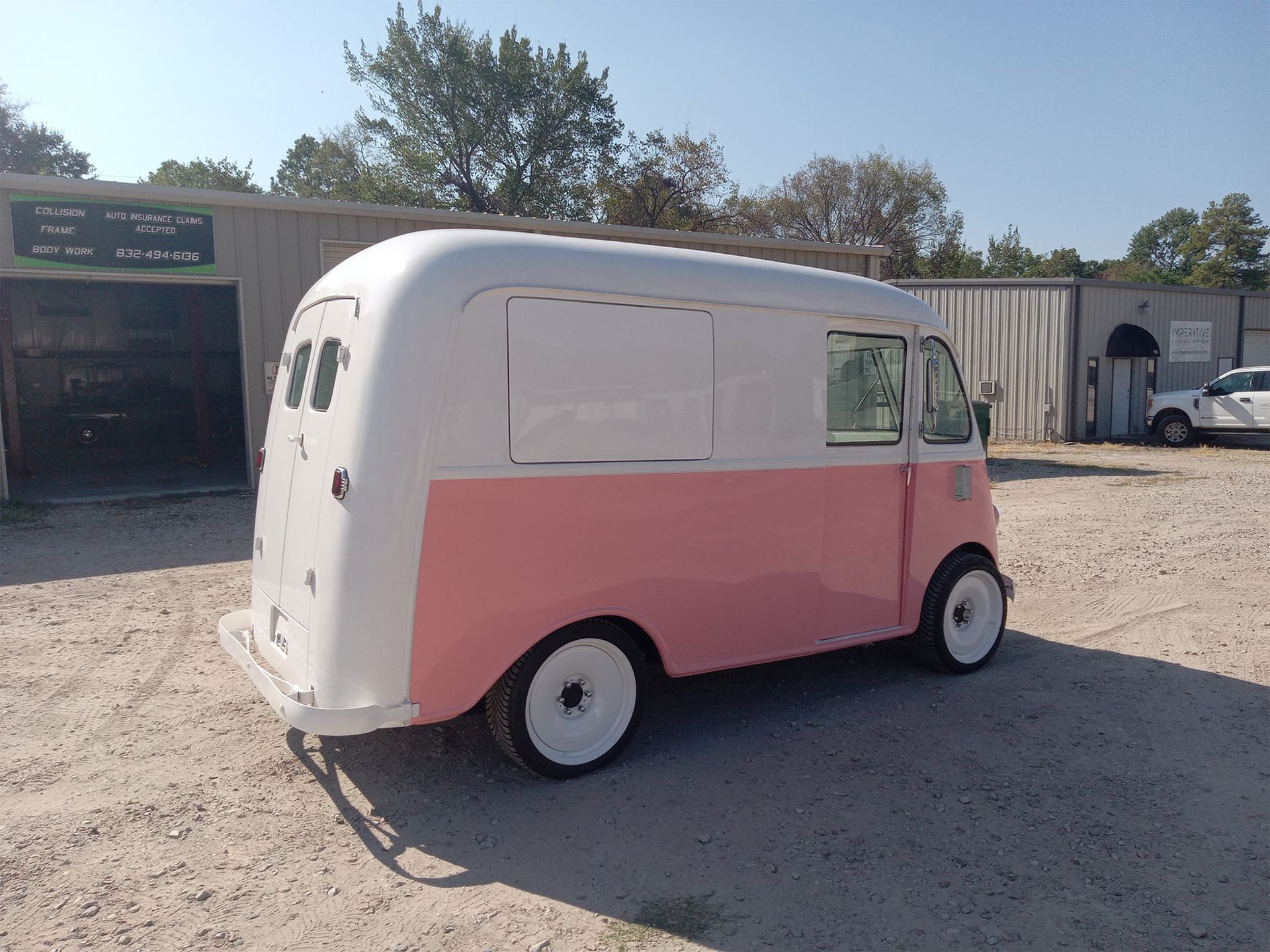 Pink and white vintage truck with large side windows, parked outside on gravel.
