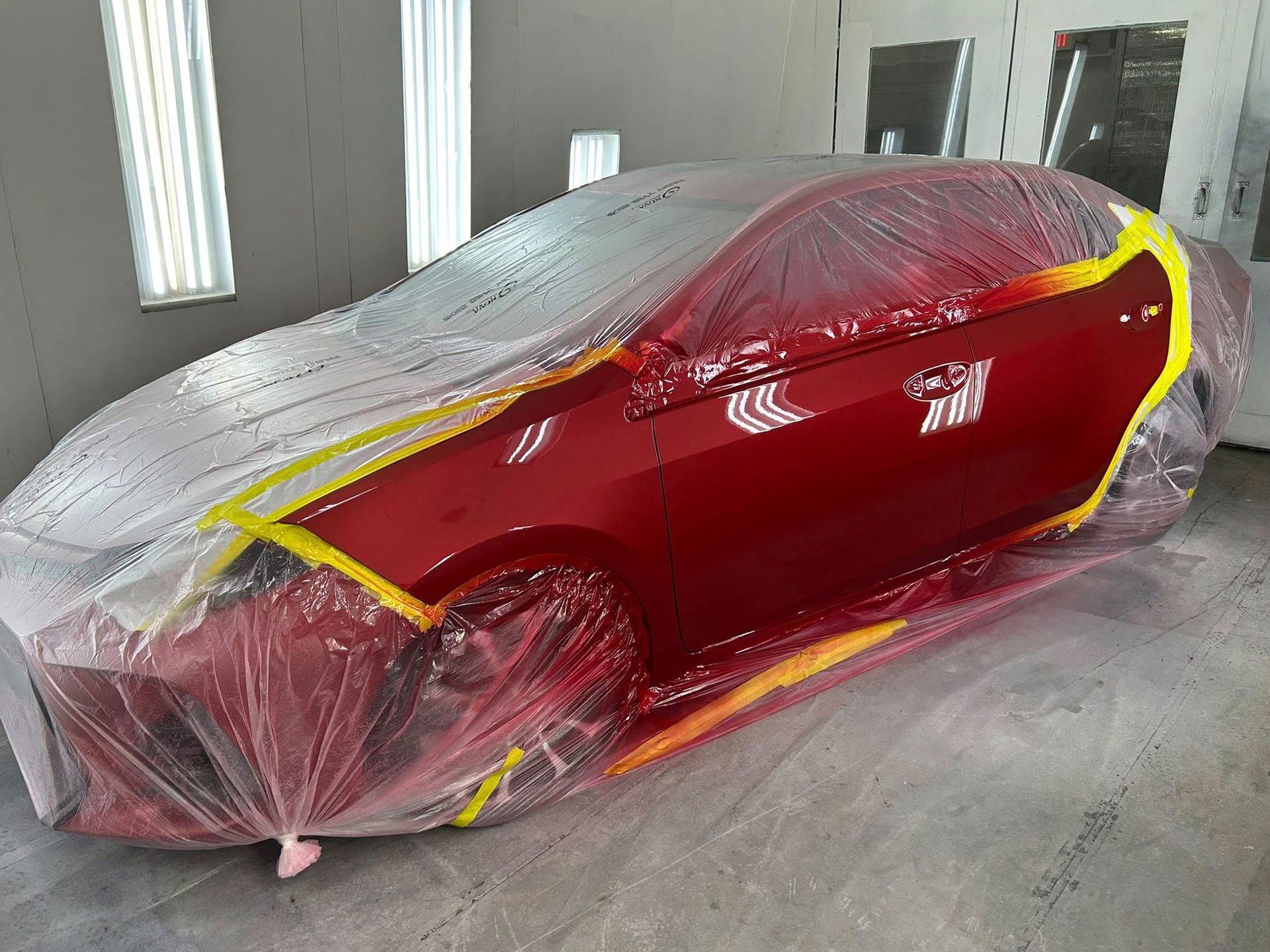 Red car in a paint booth, masked with plastic and yellow tape, ready for painting.