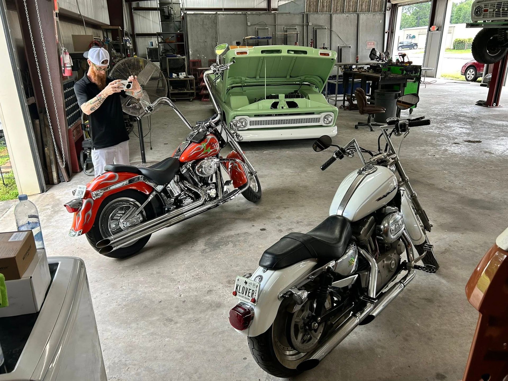 Two motorcycles in a garage, one red, one white. A man works on motorcycle parts near a green pickup.