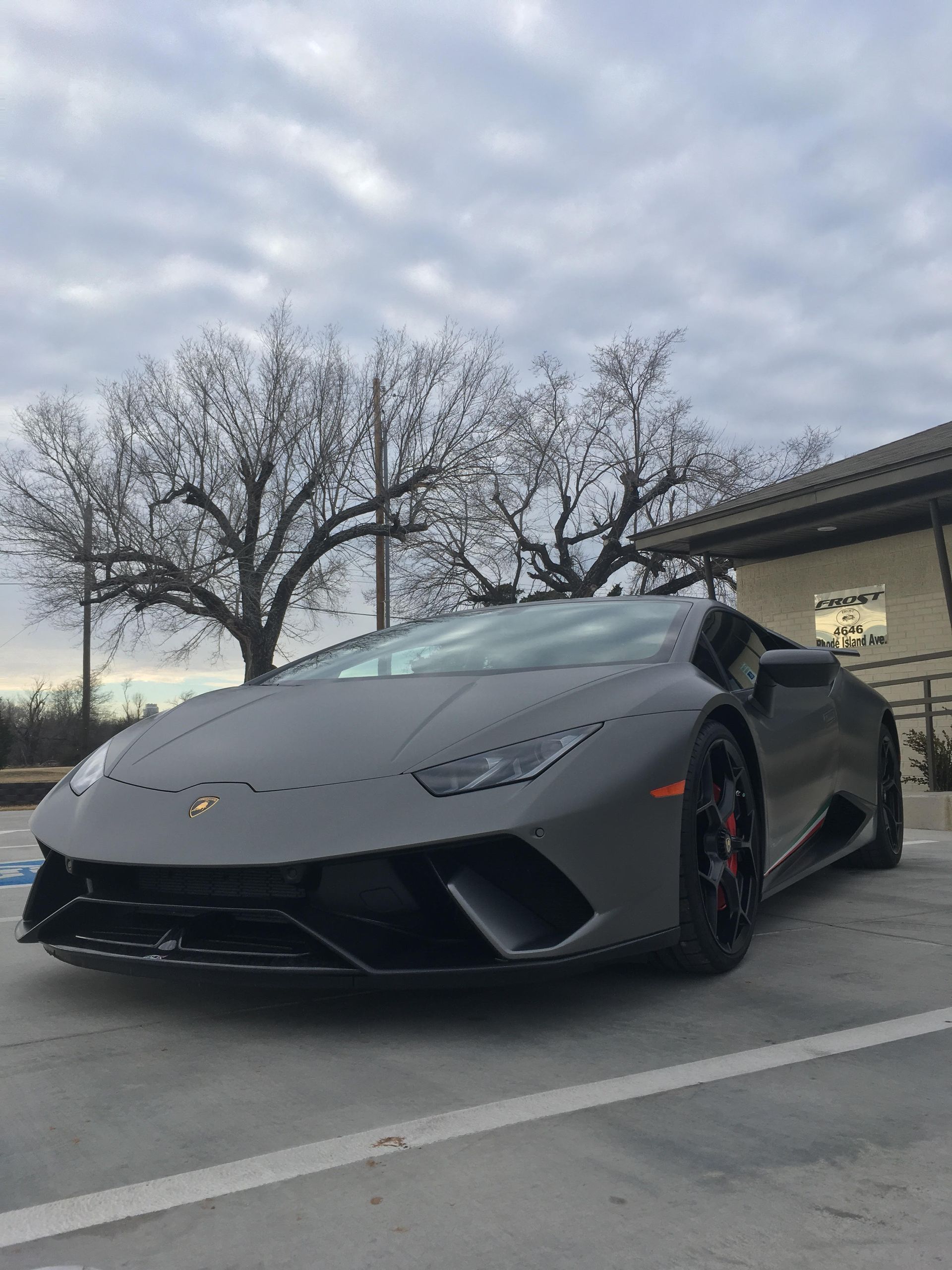 Gray Lamborghini Huracán parked in front of a building, with bare trees and a cloudy sky in the background.