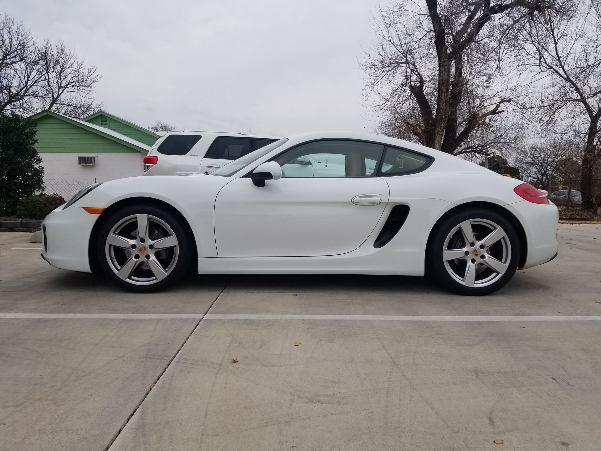 White Porsche coupe parked in a lot with a green building and white SUV in the background on a cloudy day.