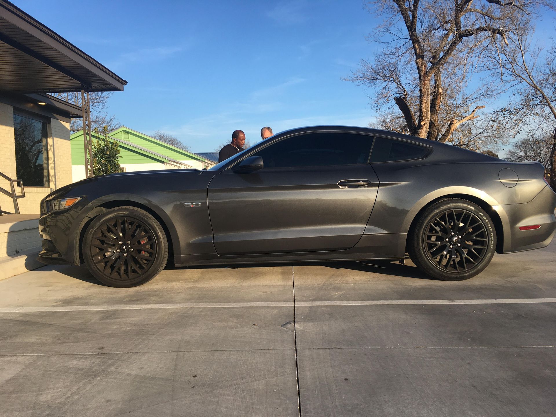 Gray Ford Mustang parked in front of a building, two people in the background.
