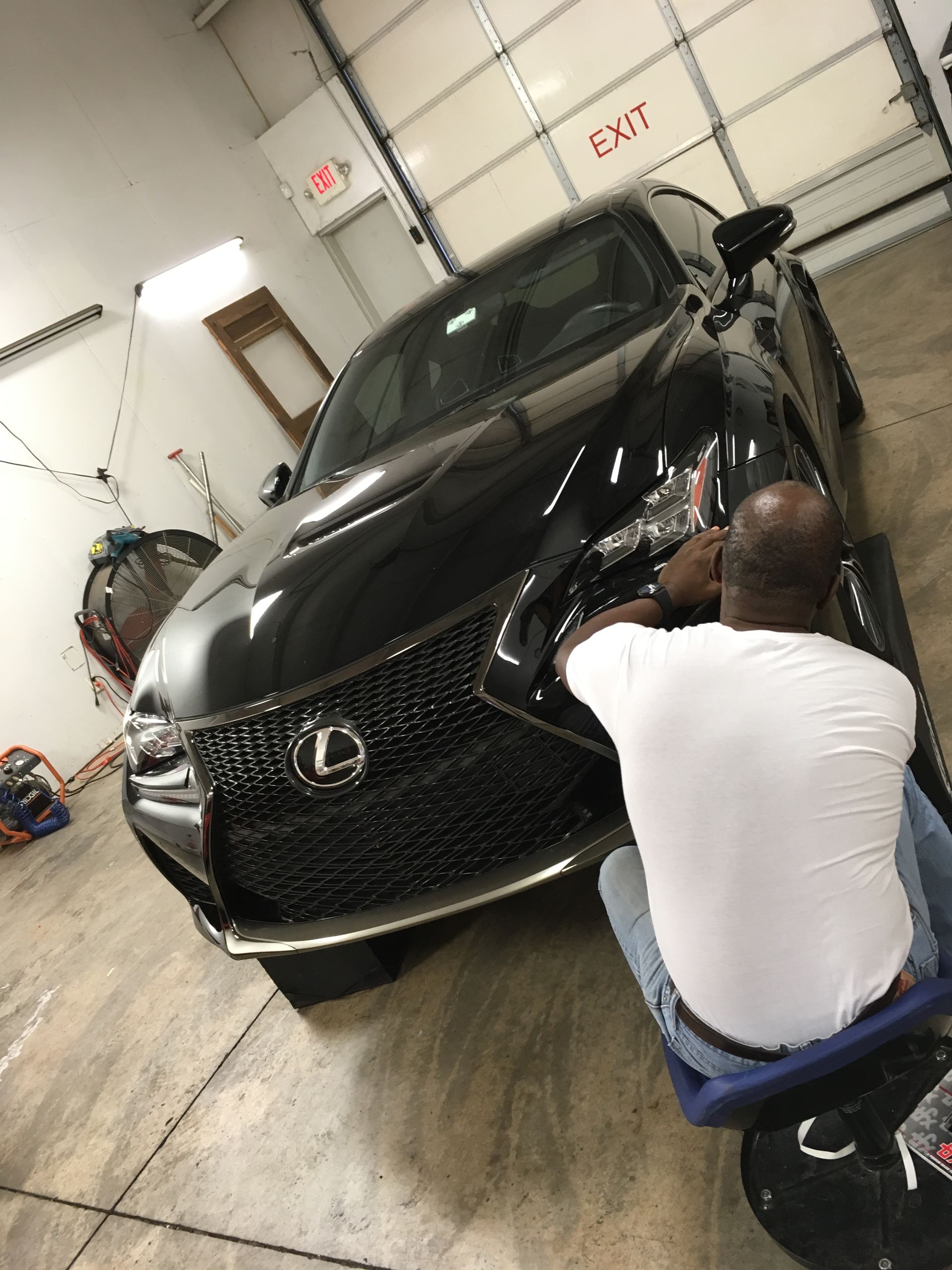 Man working on a black Lexus car in a garage; he is seated, wearing a white shirt.