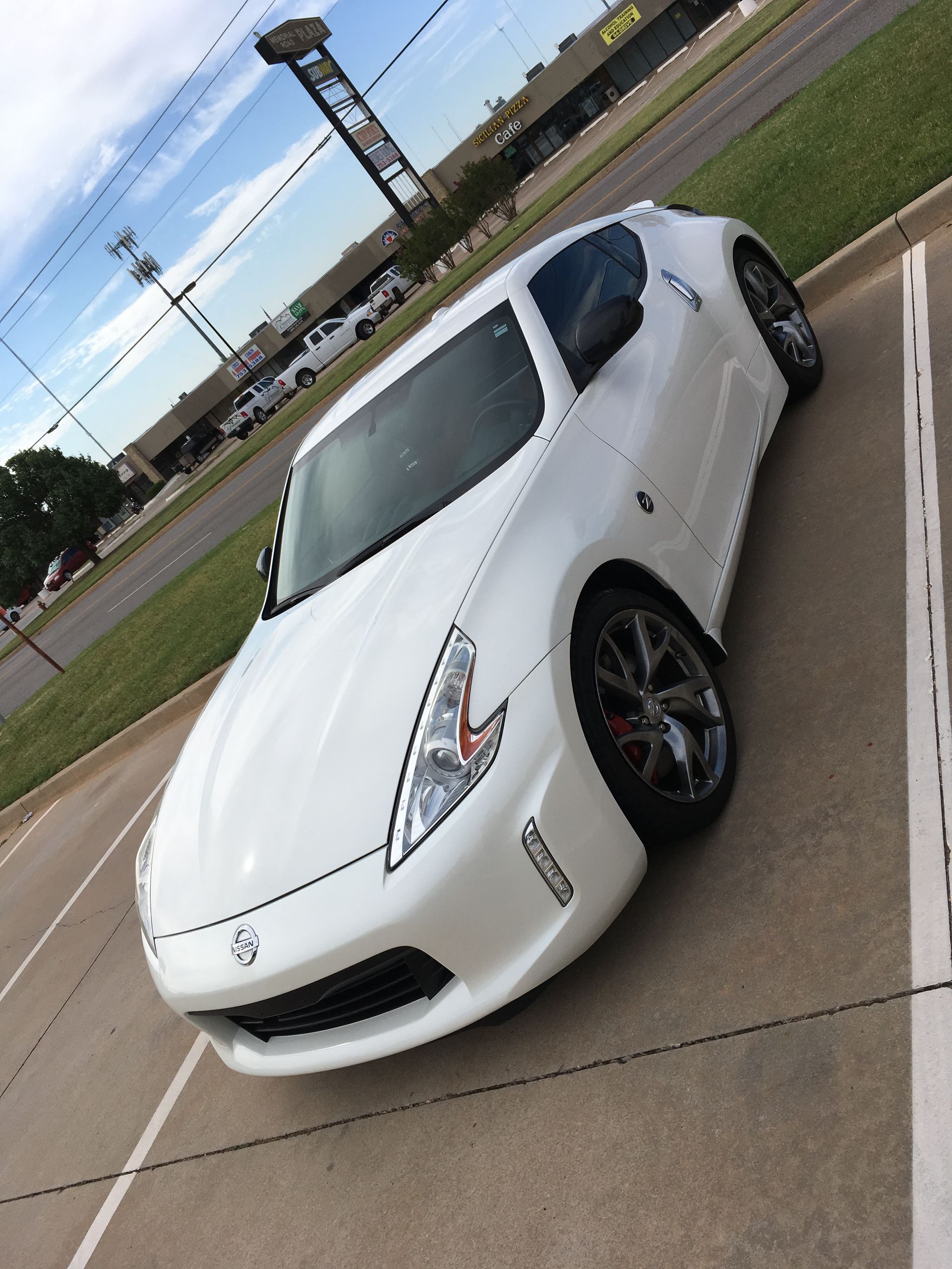 White Nissan 370Z coupe parked in a parking lot with a blue sky and buildings in the background.