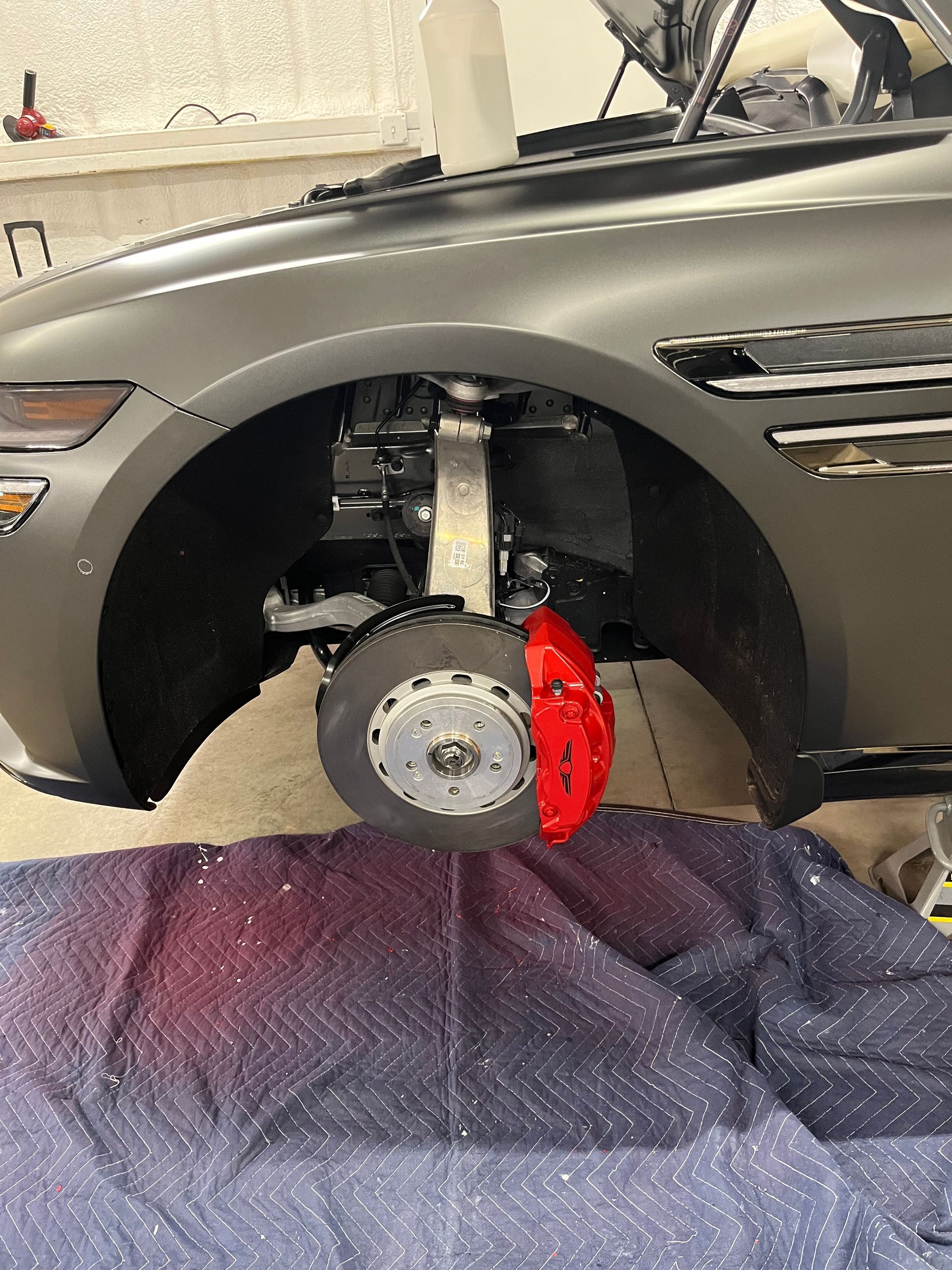 Gray car with red brake caliper; tire removed, hood open, in a garage.