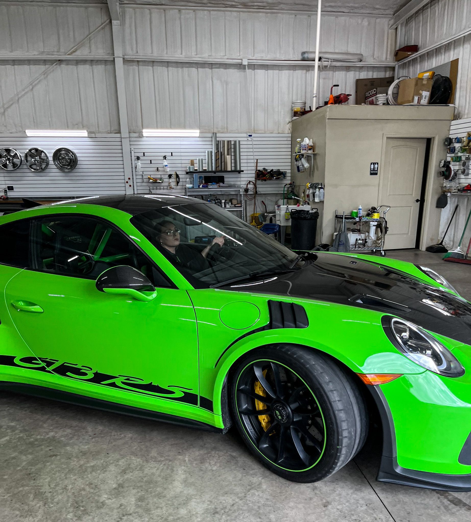 Bright green Porsche 911 GT3 RS with black hood in a garage. Driver visible.