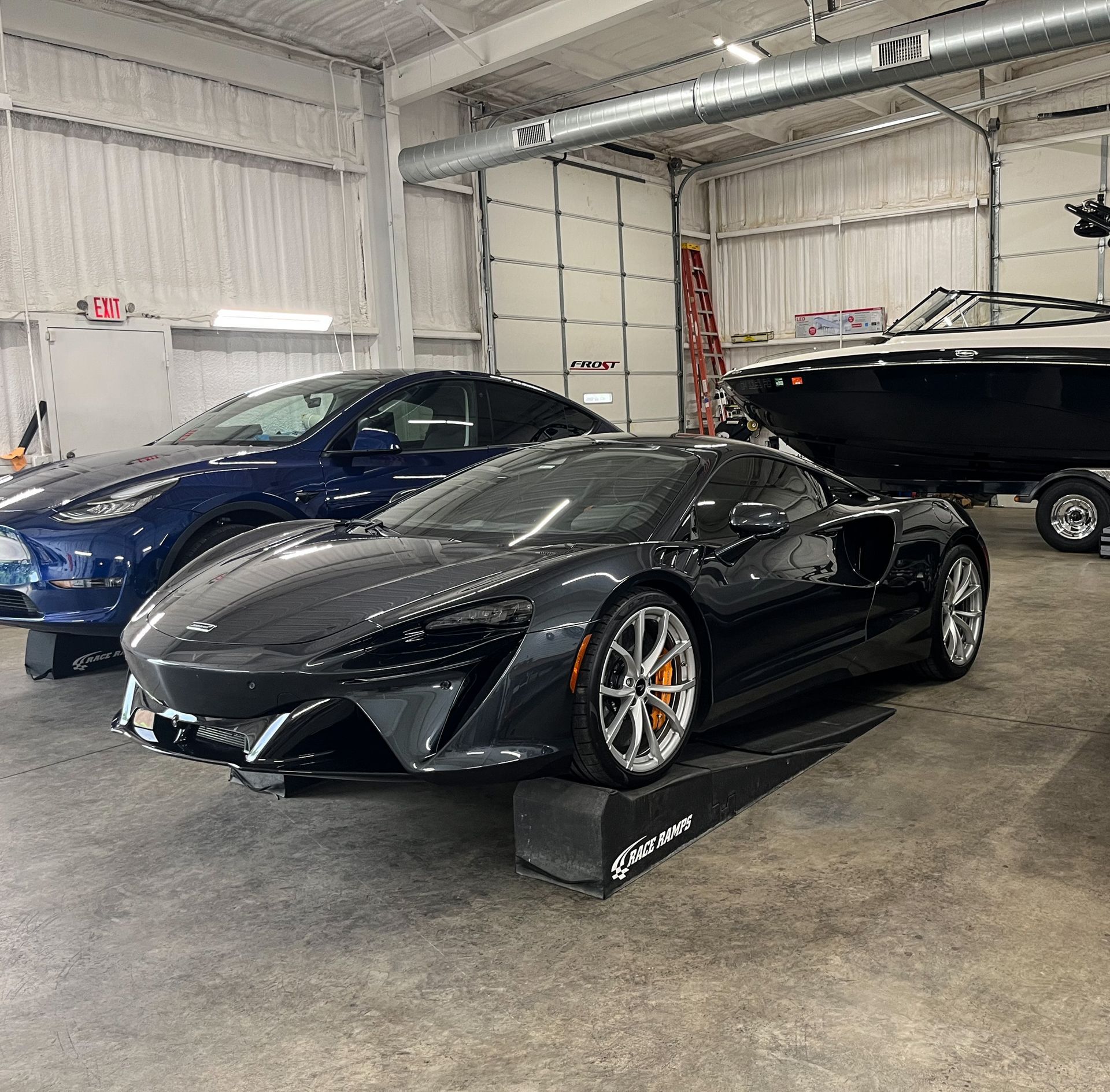 A black McLaren sports car in a garage with a blue Tesla and a boat.