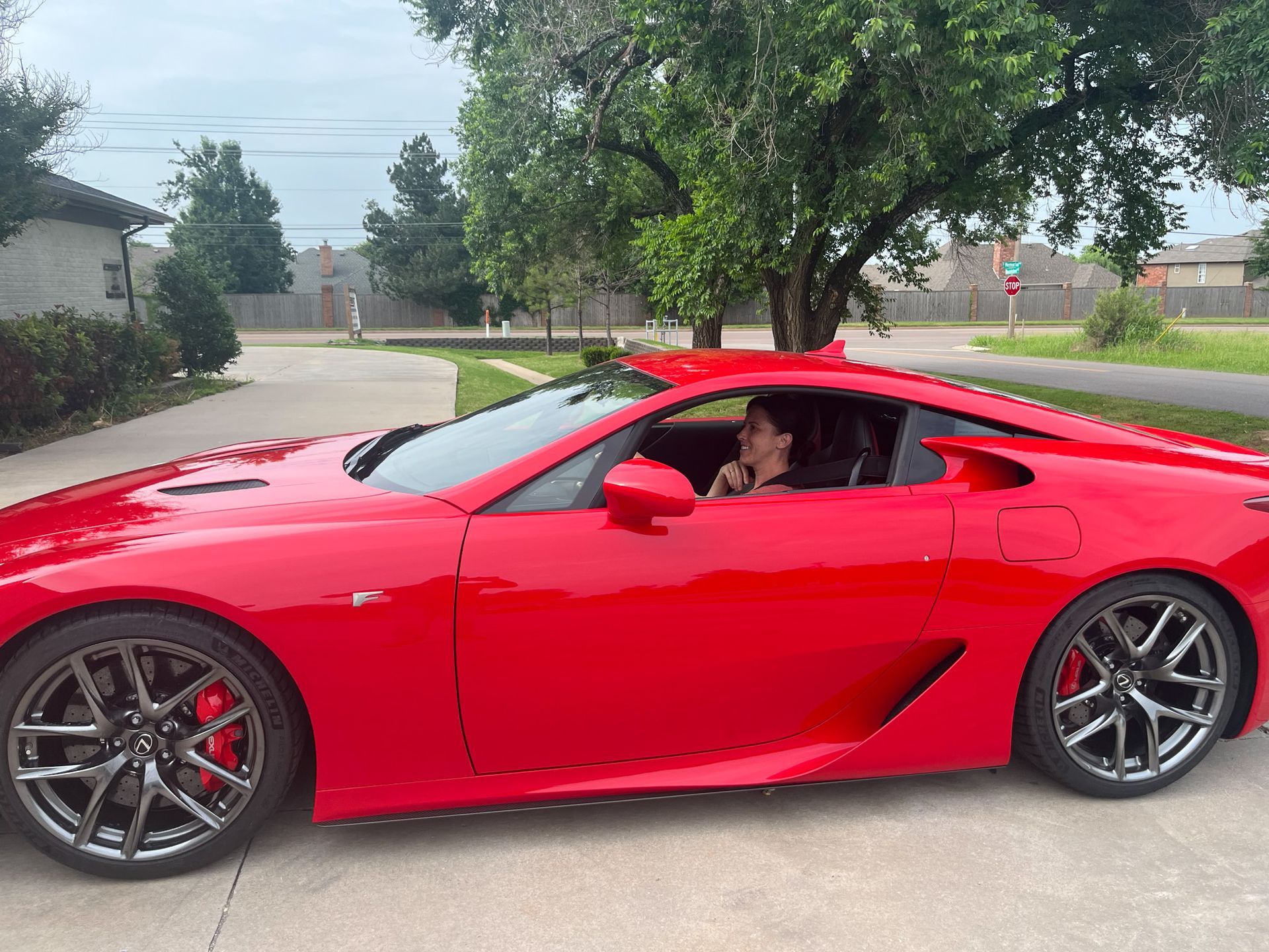 Woman in red Lexus LFA sports car on a driveway; trees and houses in the background.
