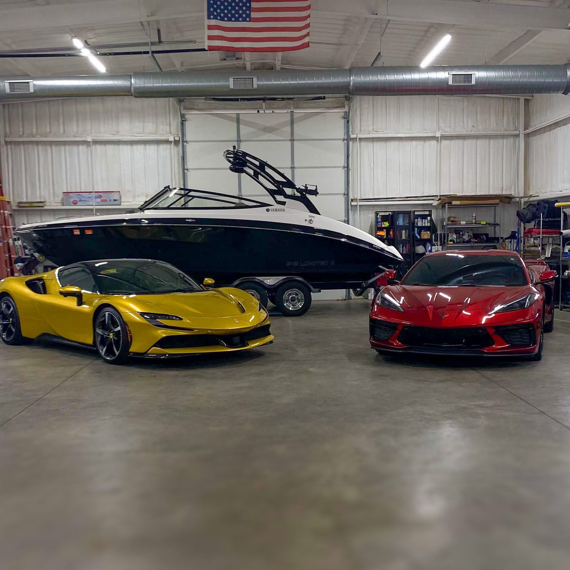 Yellow sports car and red Corvette in garage with a boat. American flag hangs above.