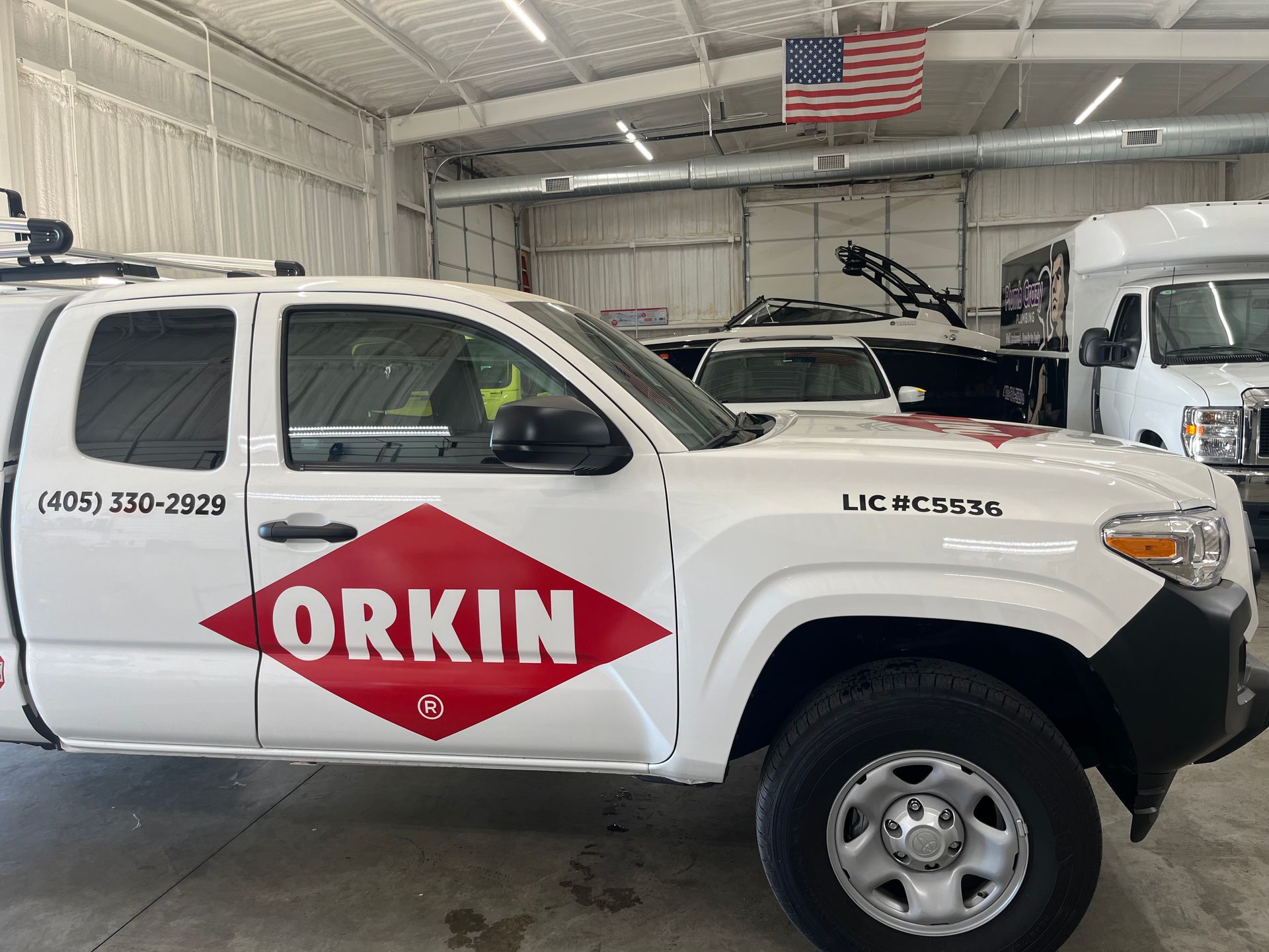 White Orkin truck parked inside a garage with other vehicles, American flag in background.