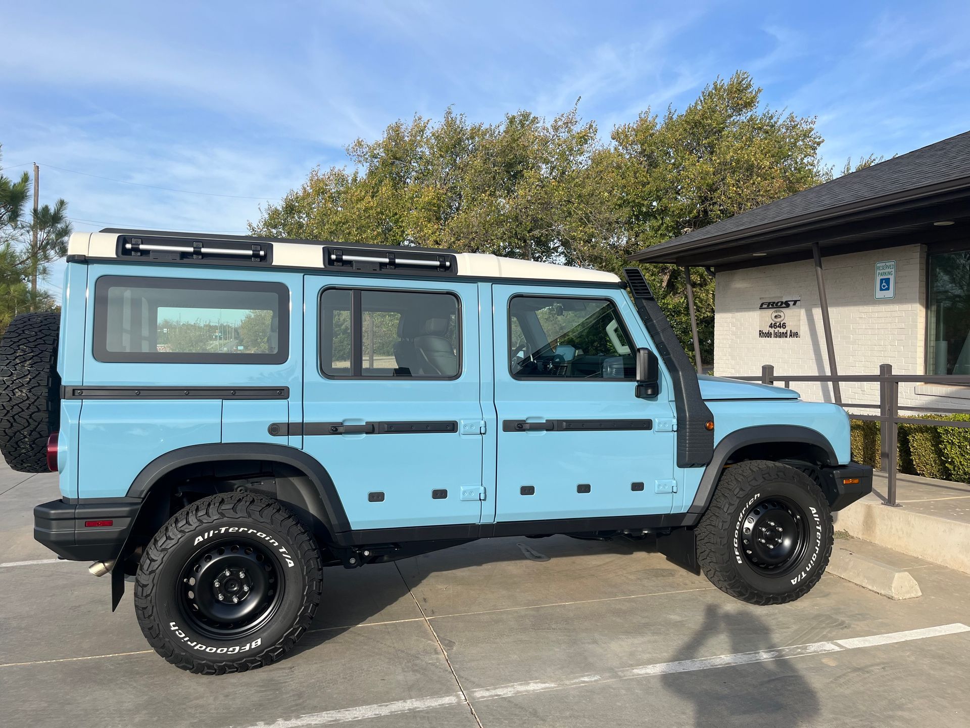 Light blue Ineos Grenadier SUV parked outside a building with a white roof, black wheels, and a spare tire.