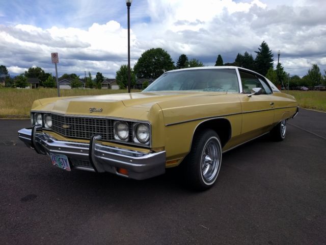 Yellow 1970s Chevrolet Impala coupe, white roof, parked outdoors on a paved surface, with a cloudy sky background.