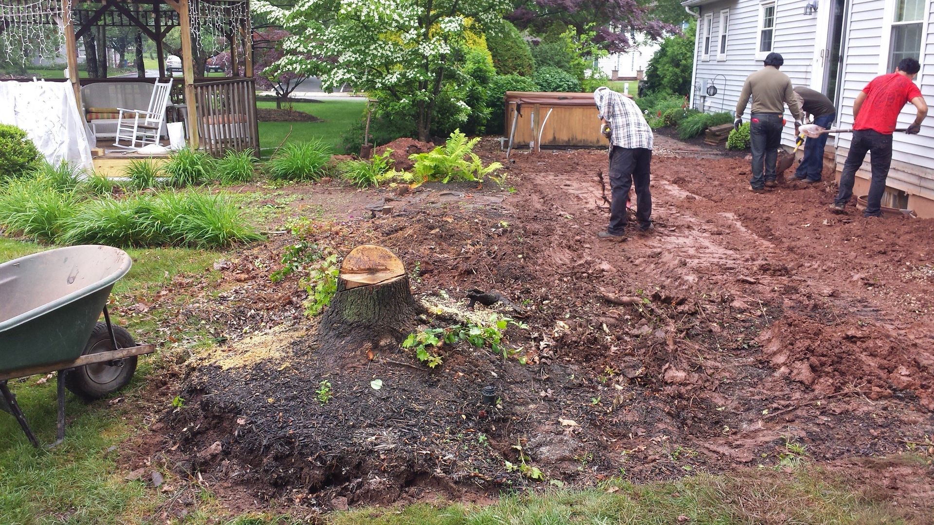 A group of people are working in a yard with a wheelbarrow in the foreground.