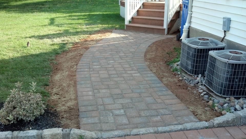 A brick walkway leading to a house with two air conditioners on the side of it.
