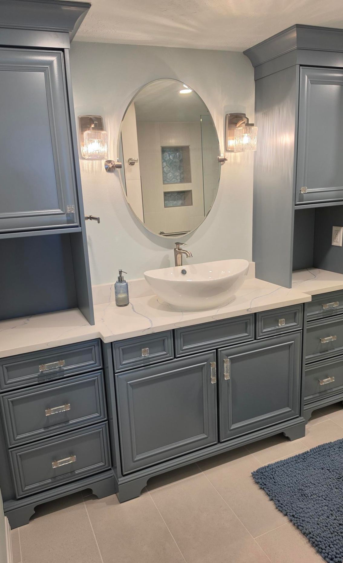 Gray bathroom vanity with oval mirror, white vessel sink, and wall sconces in a clean modern interior