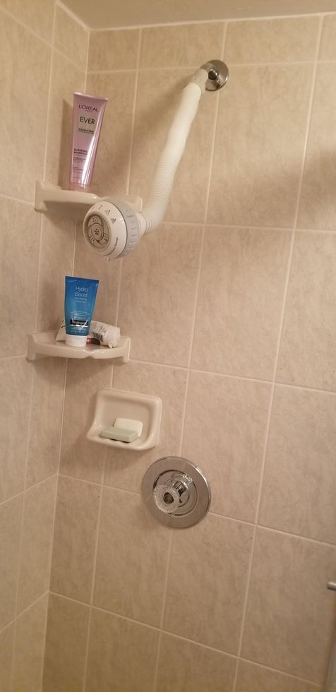Tiled shower wall with grab bar, showerhead, soap dispenser, and built-in shelf.