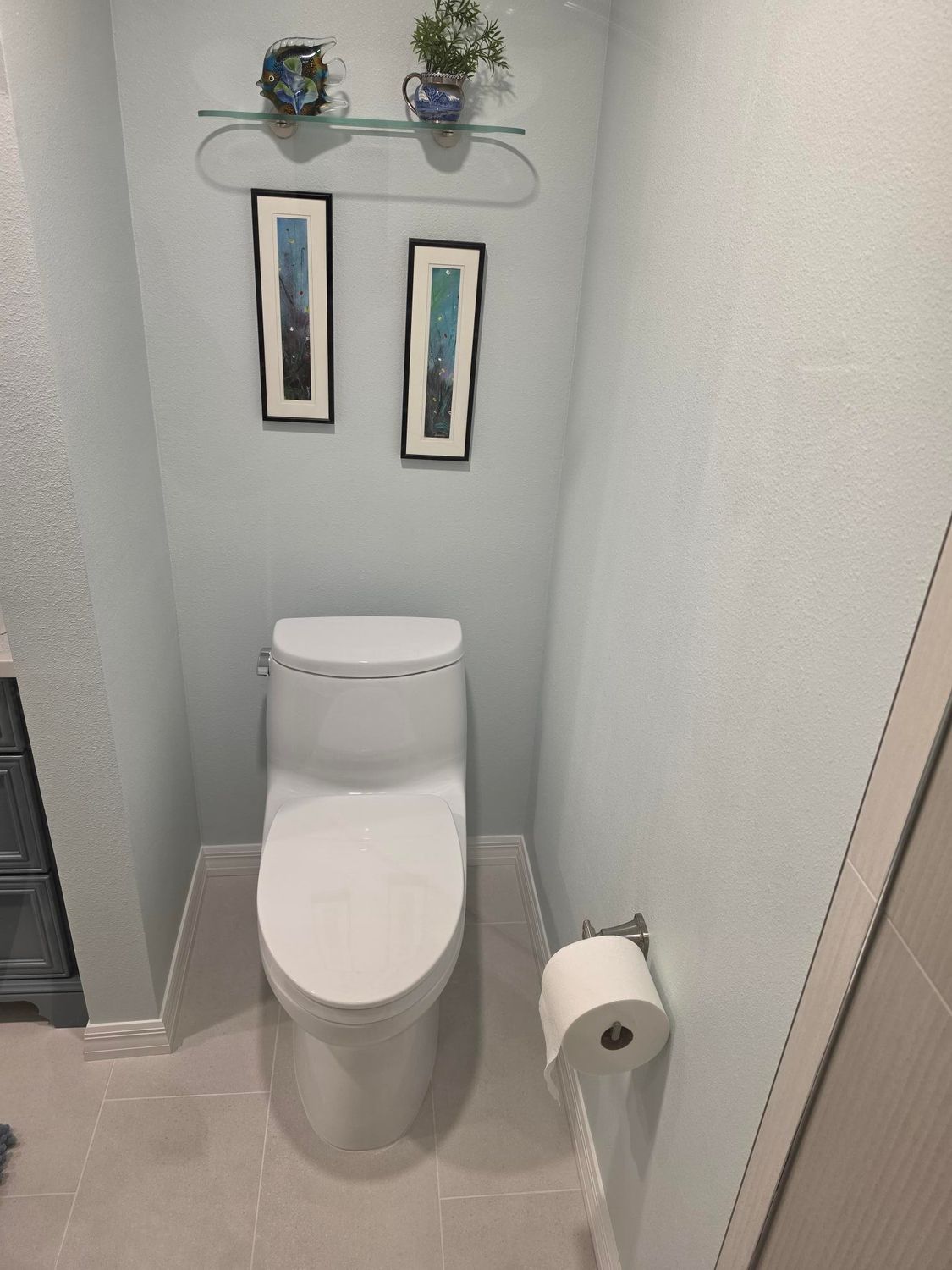 Small narrow bathroom with a toilet, toilet paper roll, wall shelf, and two framed pictures.