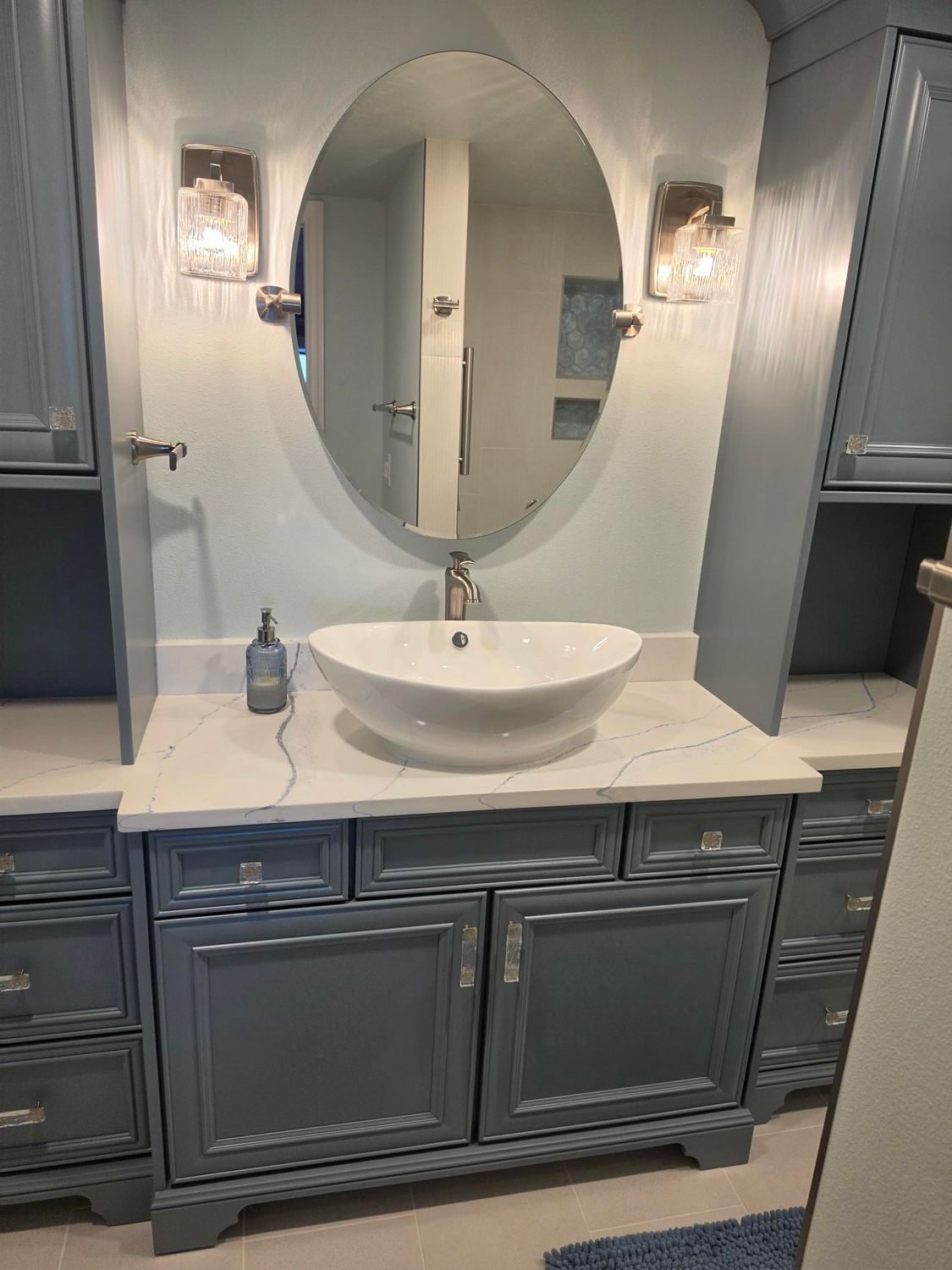 Gray bathroom vanity with oval mirror, white vessel sink, and wall sconces in a clean modern interior