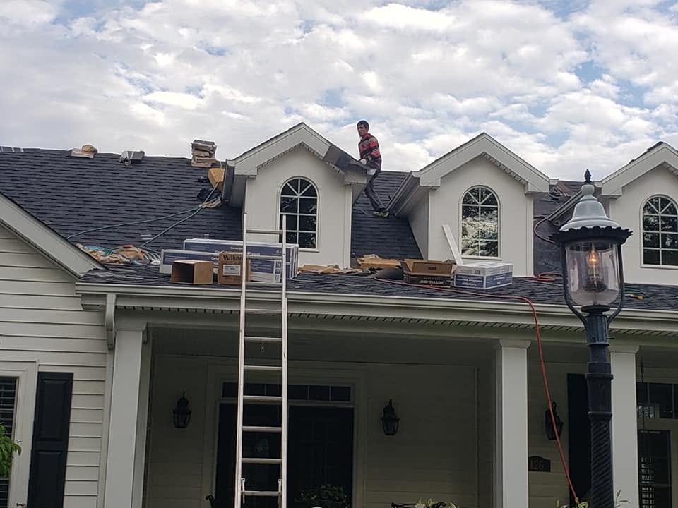 A worker repairs the roof of a house with several gabled dormers on a bright, cloudy day.