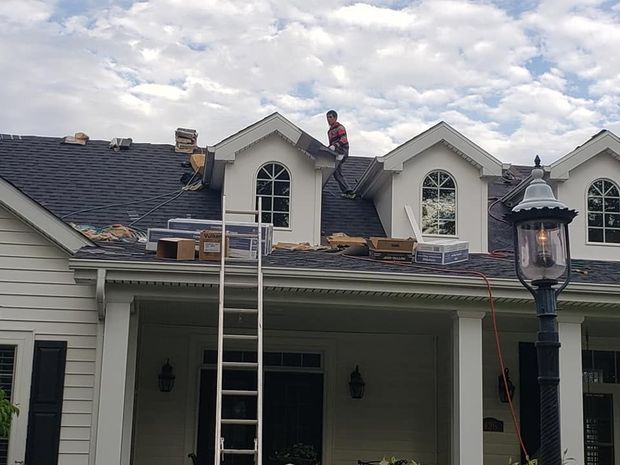 A worker repairs the roof of a house with several gabled dormers on a bright, cloudy day.